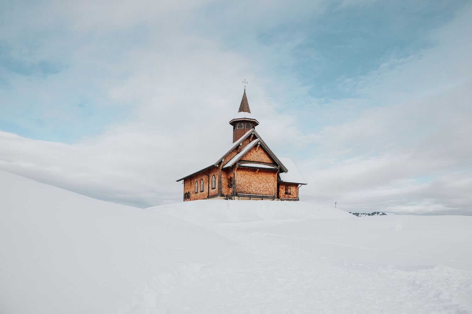 Rustic wooden chapel nestled in pristine white snow against cloudy sky.