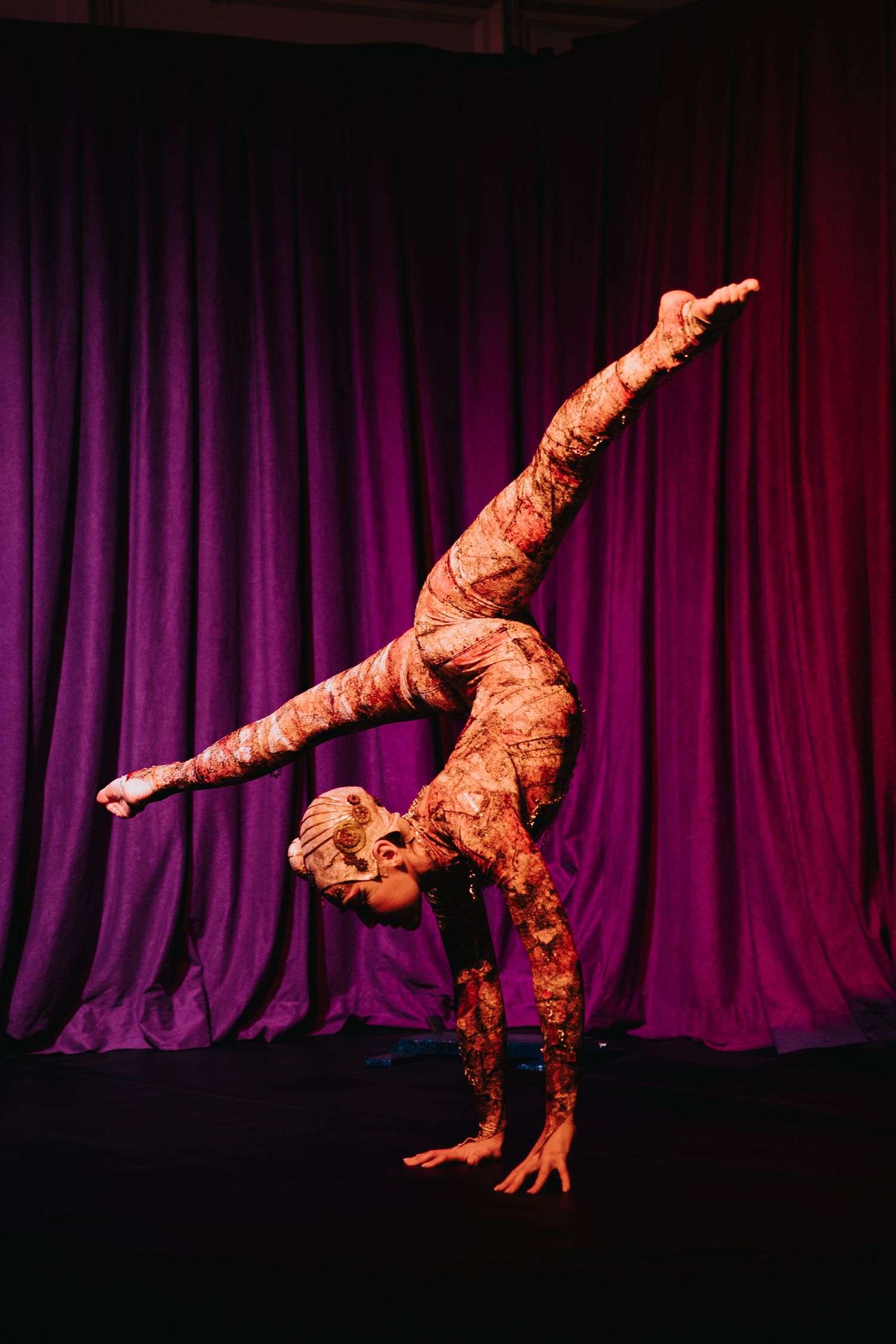 Acrobatic performer in ornate costume executing dynamic moves against a purple curtain backdrop.
