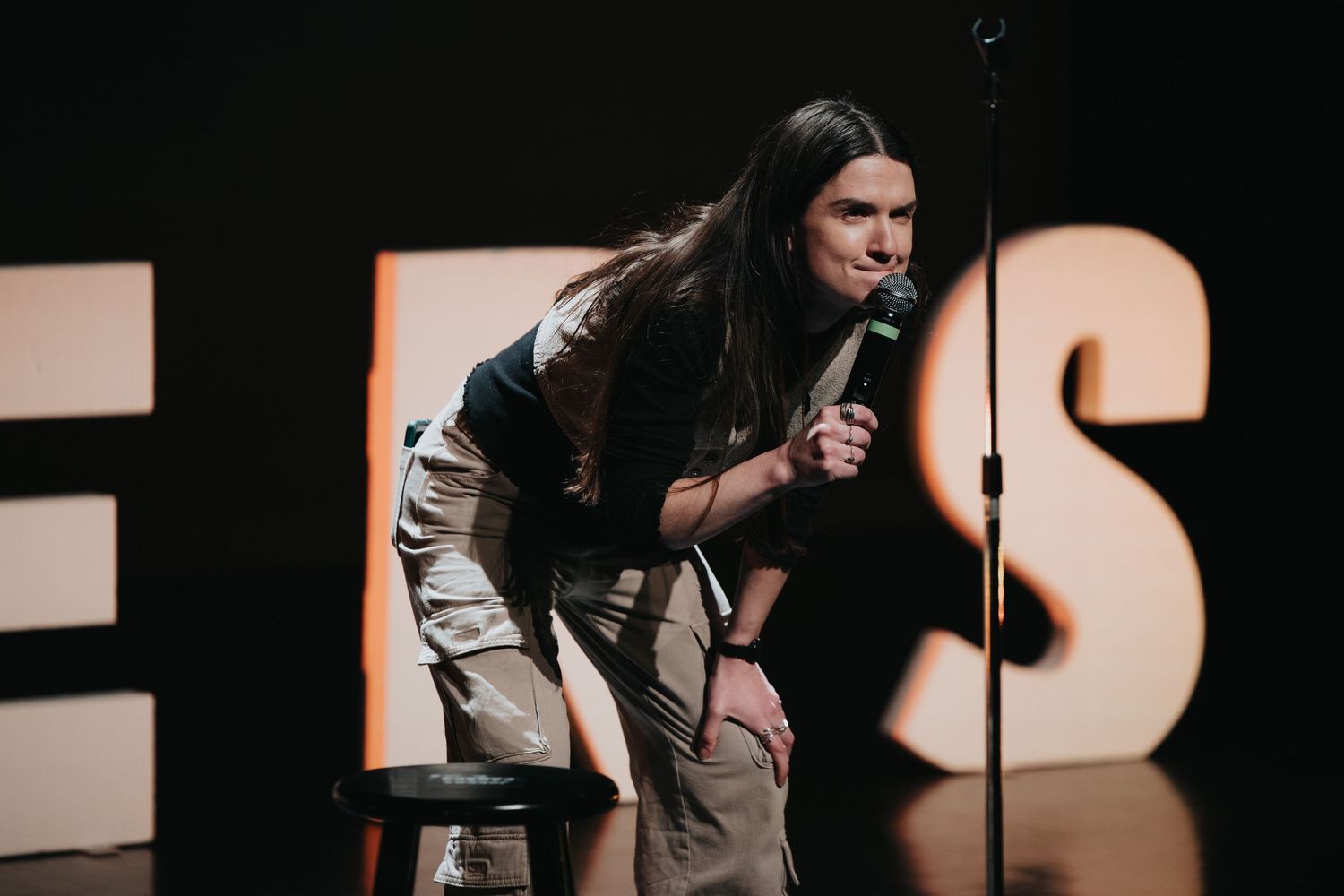 Long-haired comedian leans forward holding microphone on dimly lit stage with bold letter backdrop.
