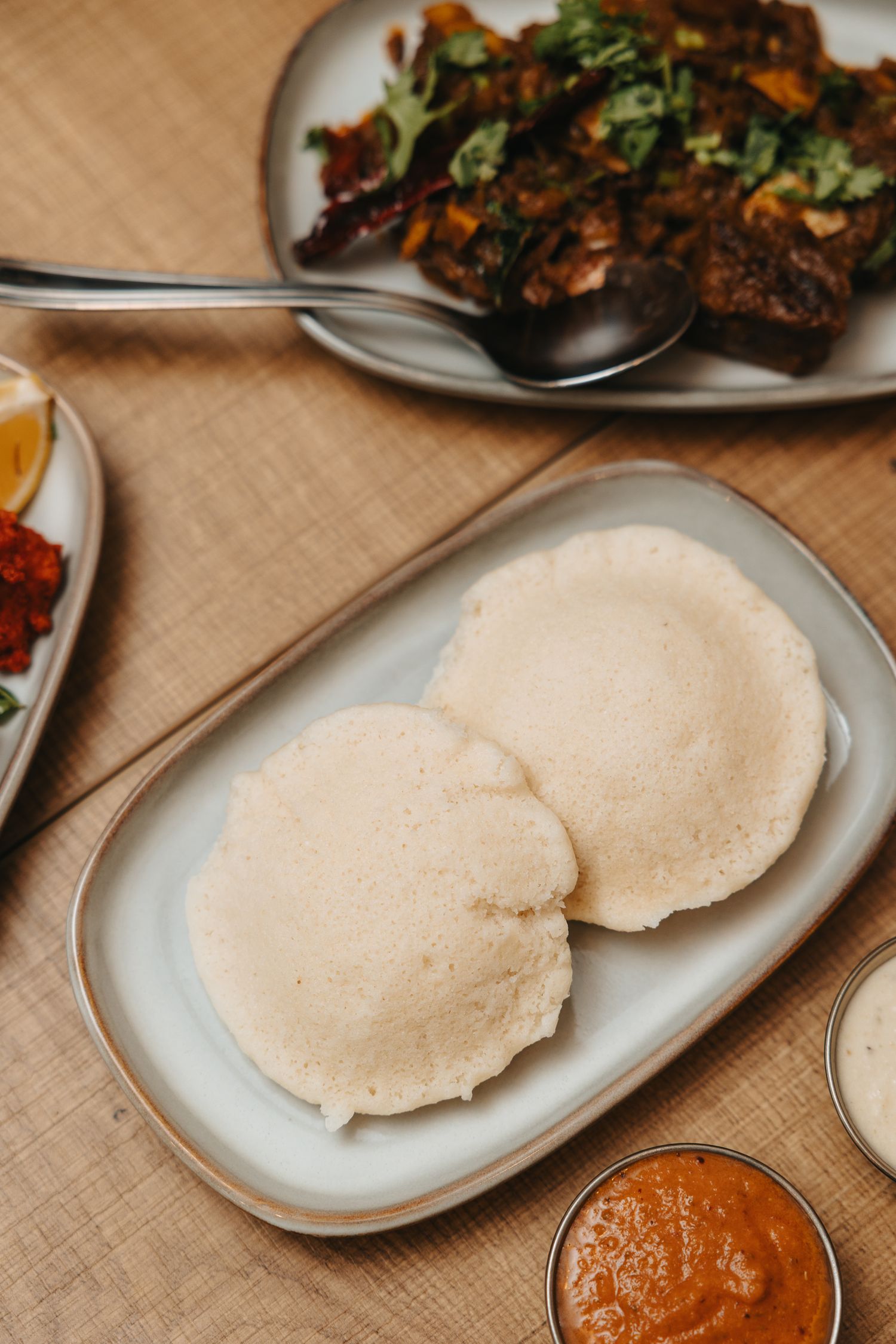 Two fluffy idli served on a ceramic plate with sambar and chutney dipping sauces, alongside spiced dishes on a wooden table.