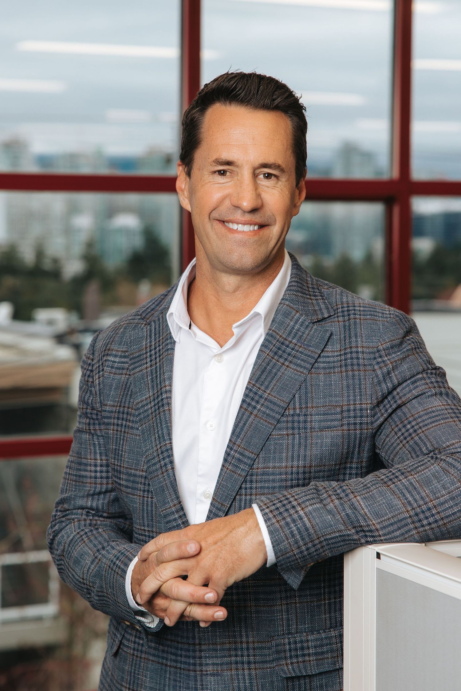 Professional wearing a gray plaid blazer and white shirt poses in front of large windows in an office setting.