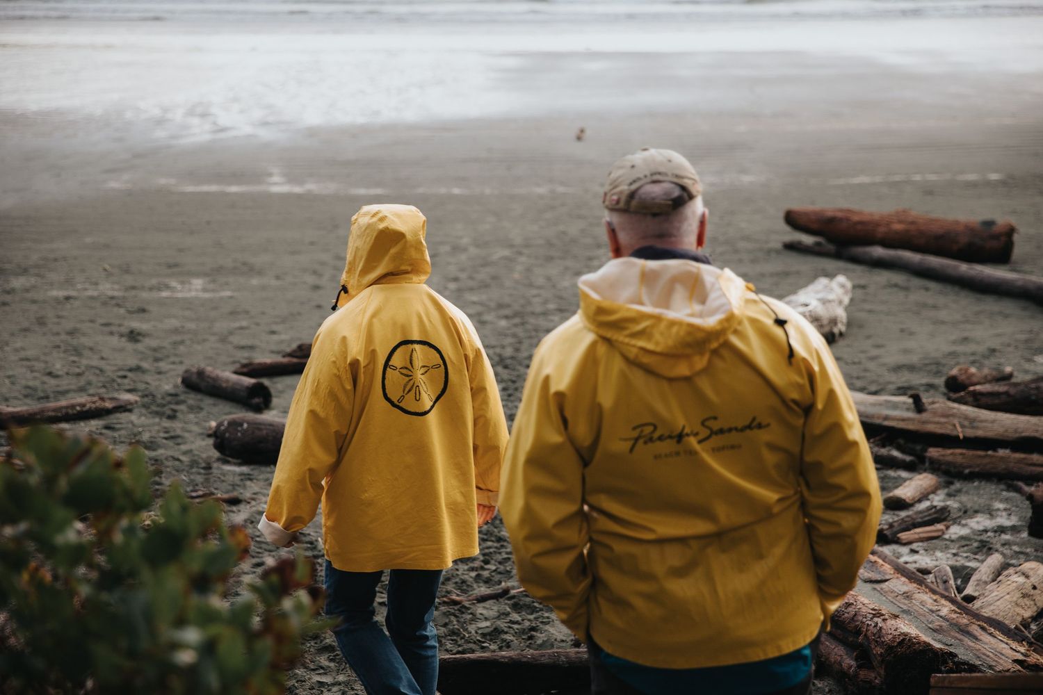 Two people in yellow jackets walking along driftwood-covered beach.