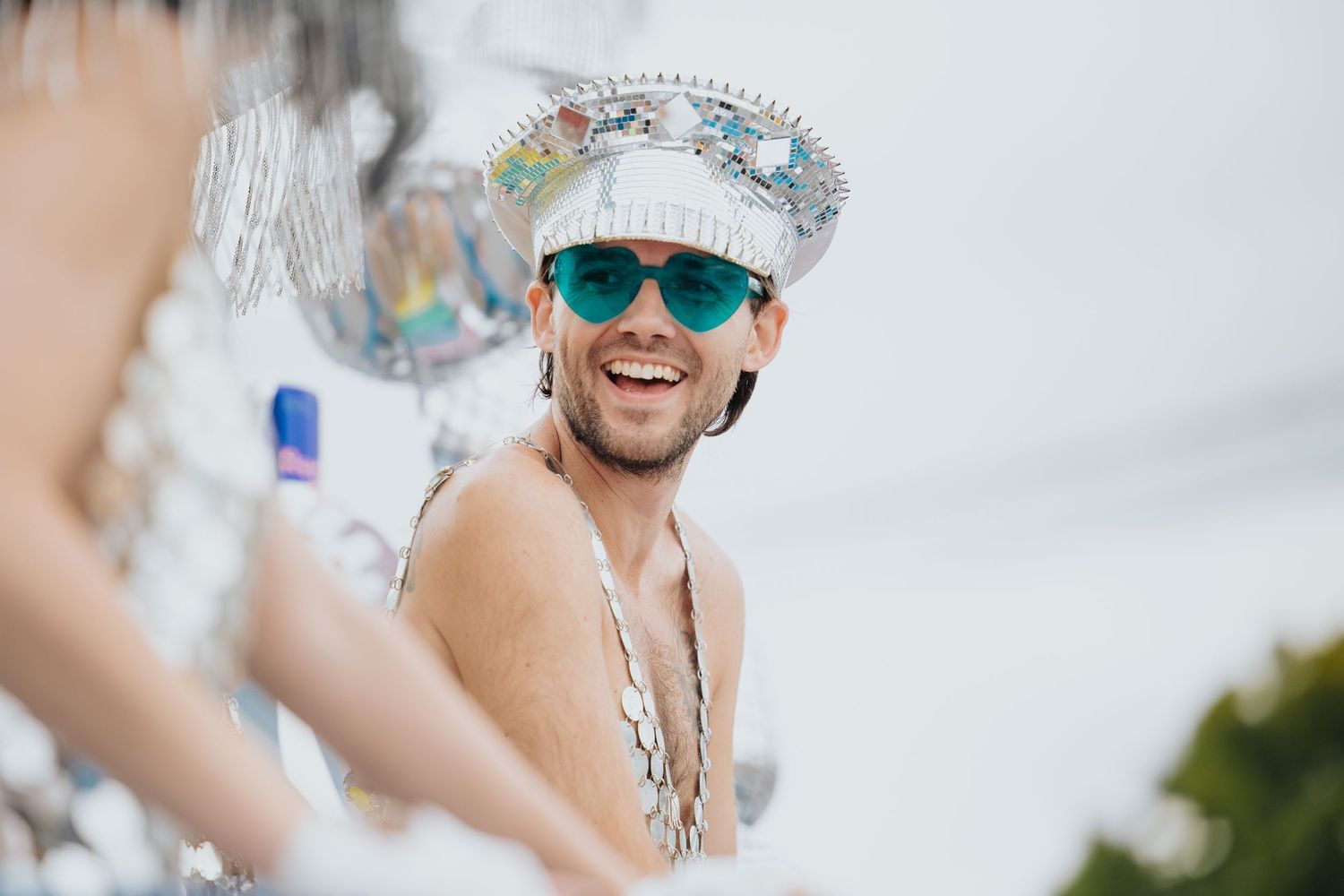 Smiling man in silver carnival costume and teal sunglasses enjoys festive outdoor celebration.