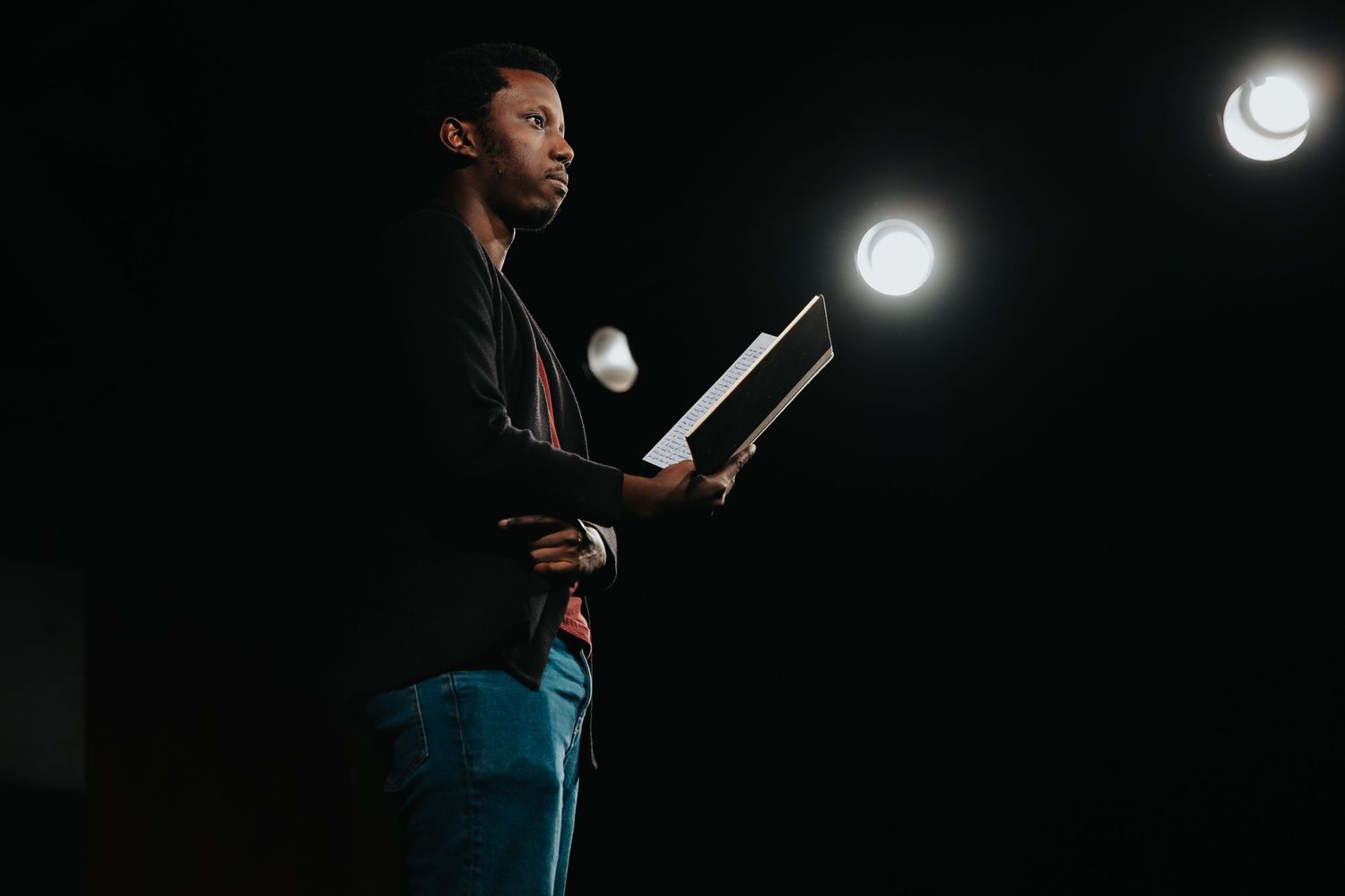 Man in black jacket reads from a binder on a dark stage with spotlights behind him.
