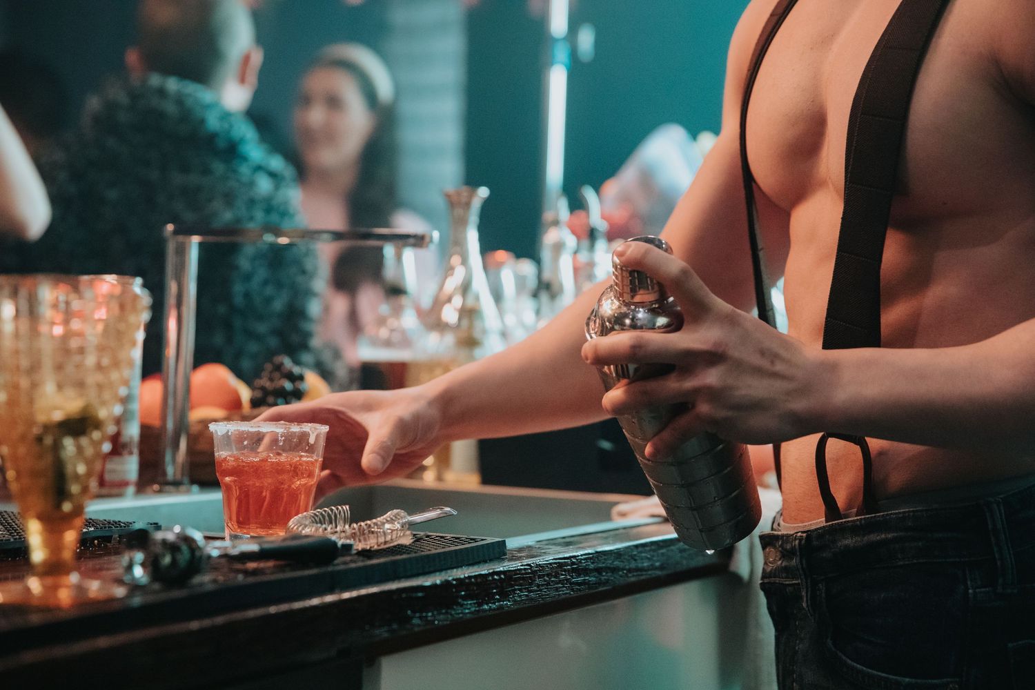 Shirtless bartender with suspenders mixing cocktails at a busy nightclub bar.