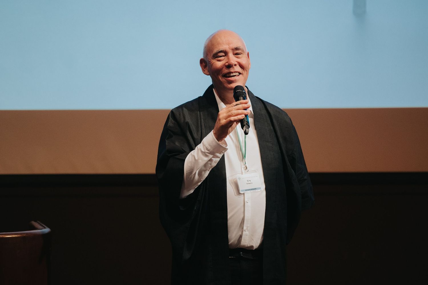 Speaker presenting on stage with professional lighting and projection screen.