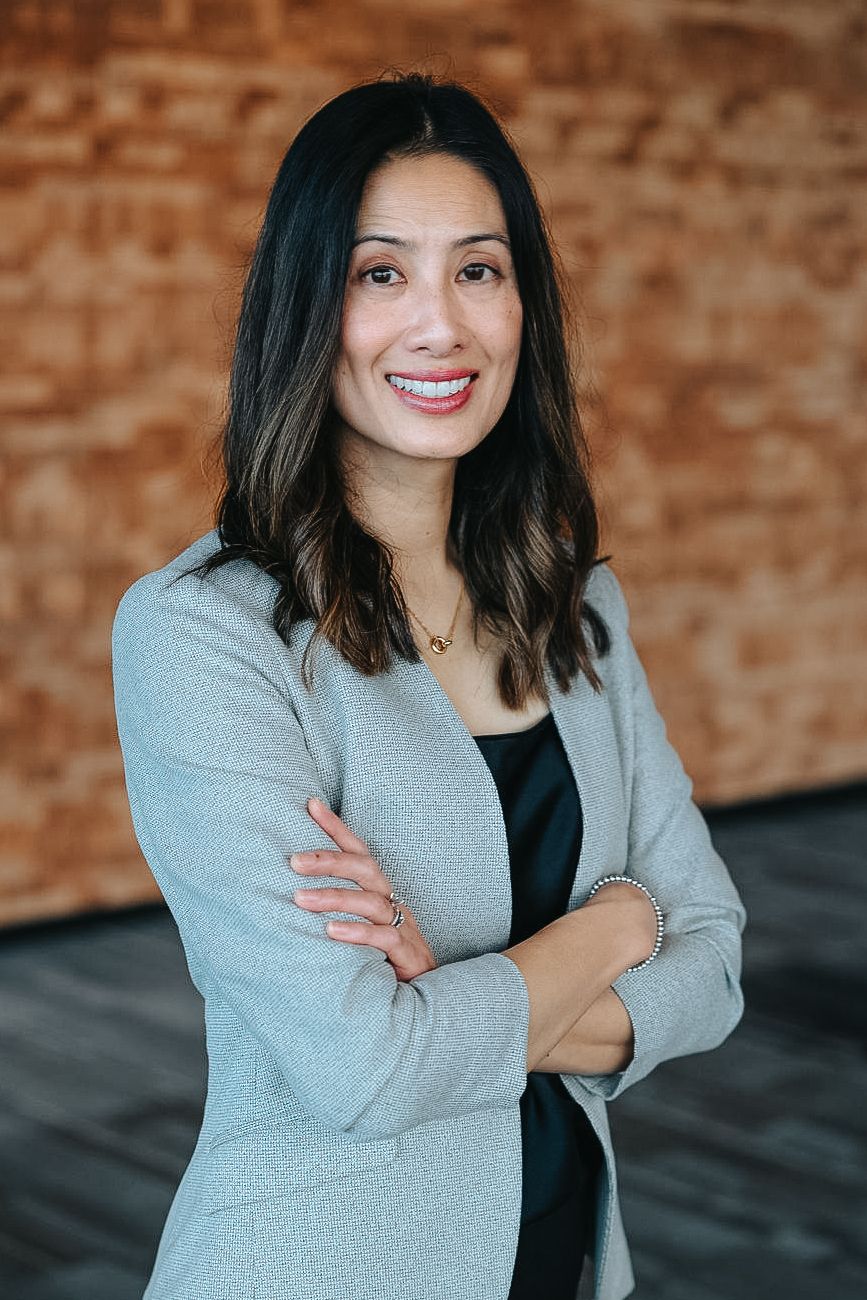 Professional headshots against a brick wall showing a person in a gray blazer and black top.