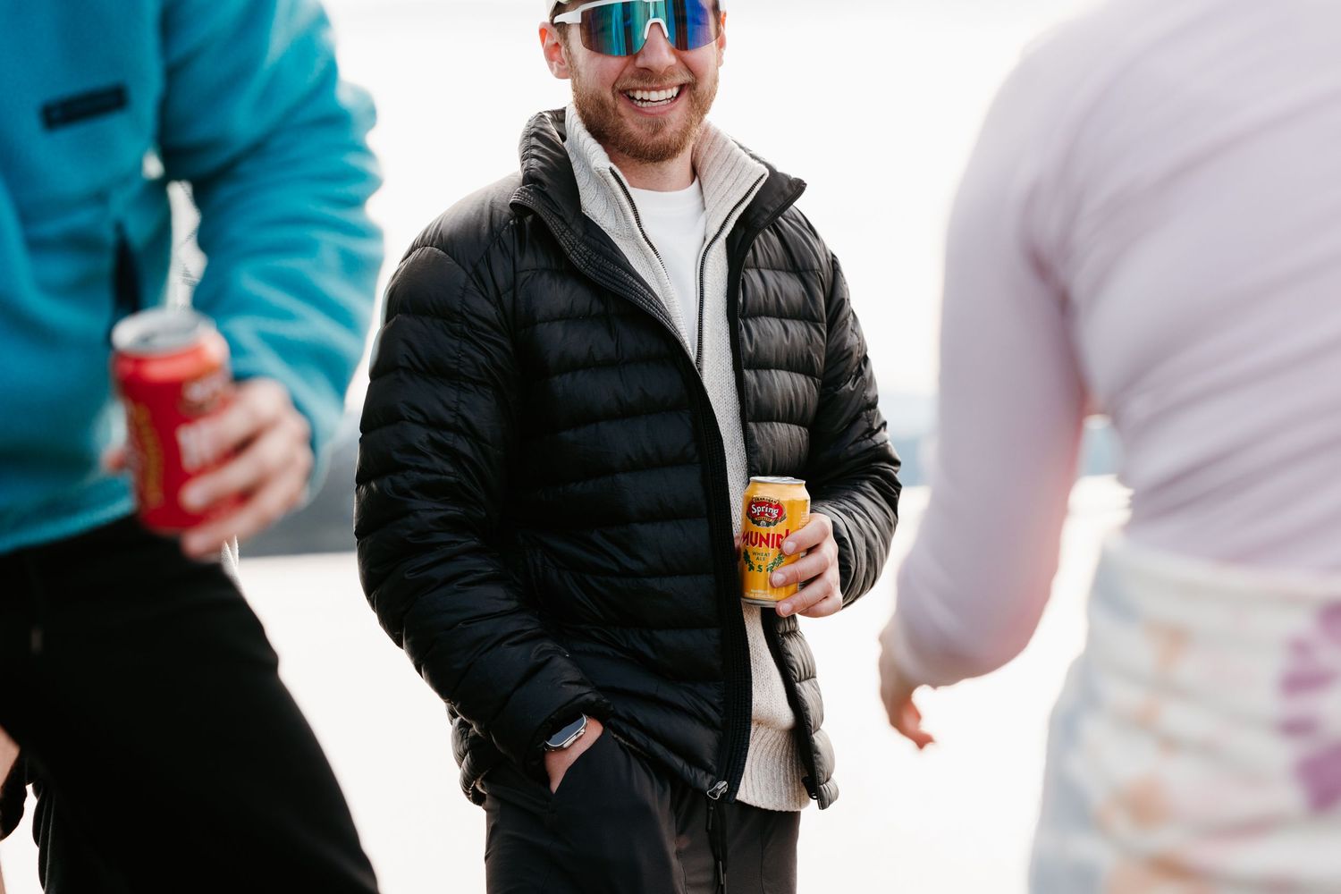 Person in sunglasses and winter jacket holding a beverage outdoors.