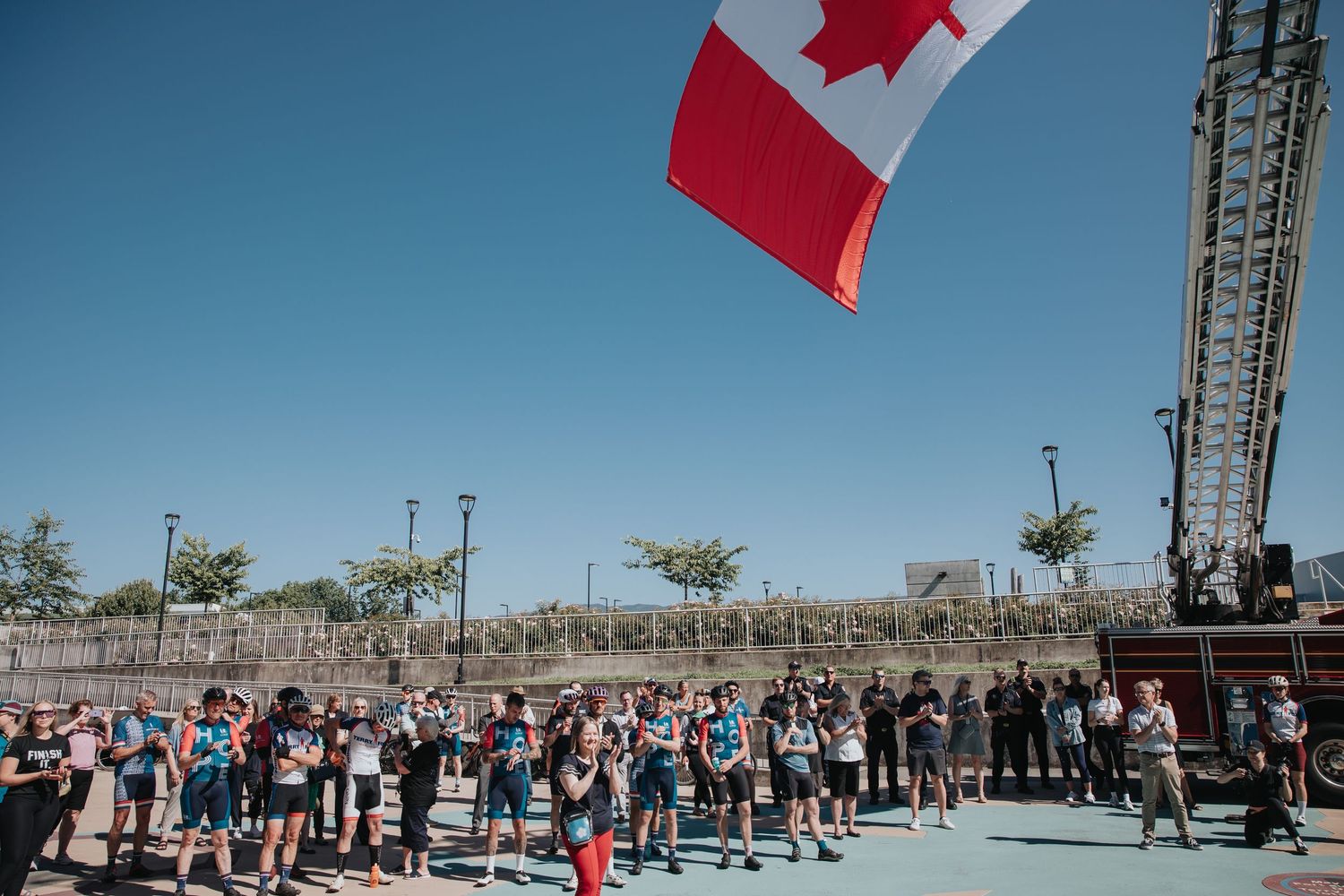 Crowd of people gathered outdoors under Canadian flags on a sunny day.
