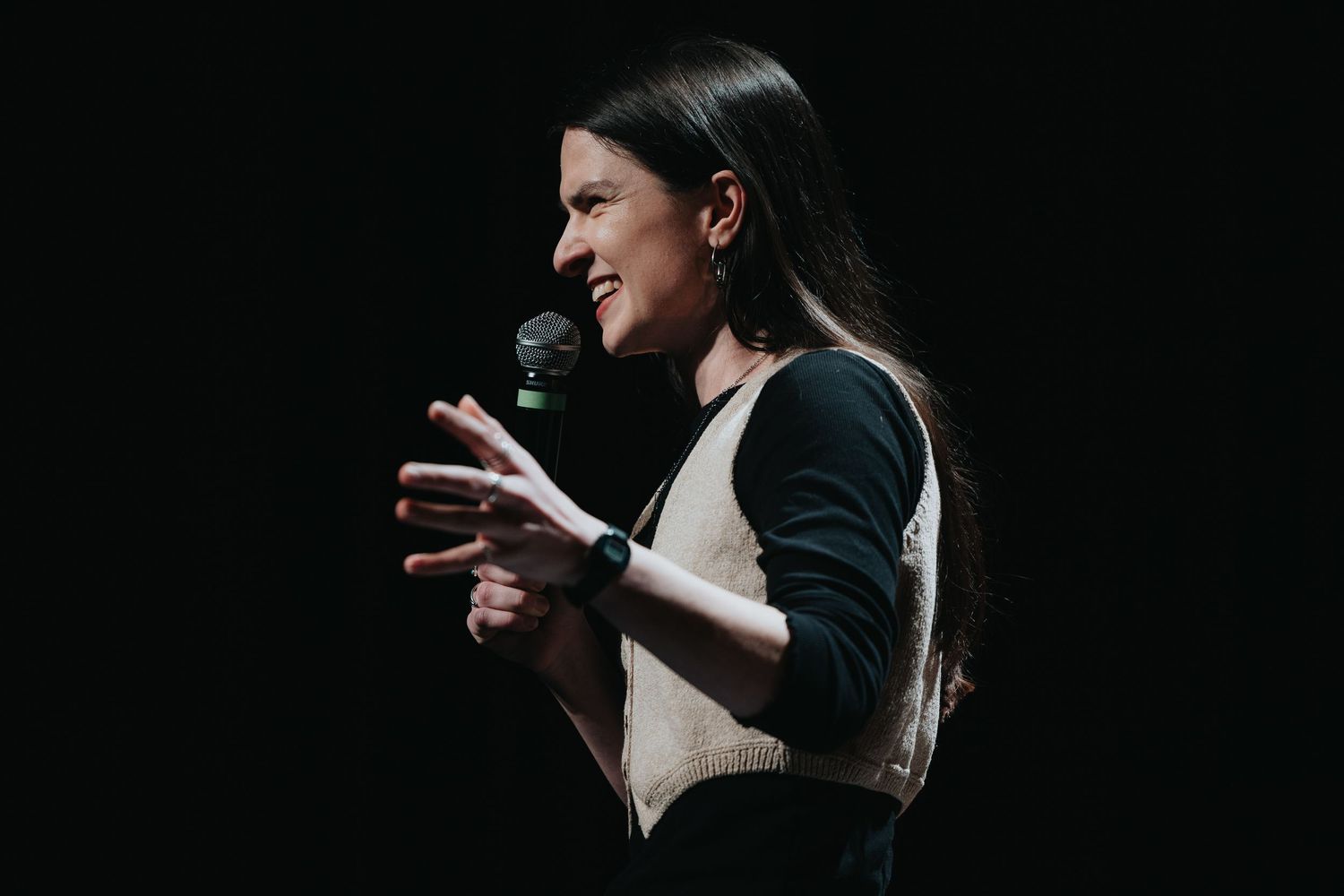 Woman speaking on stage holding a microphone, gesturing with hand, wearing a beige vest over black top.