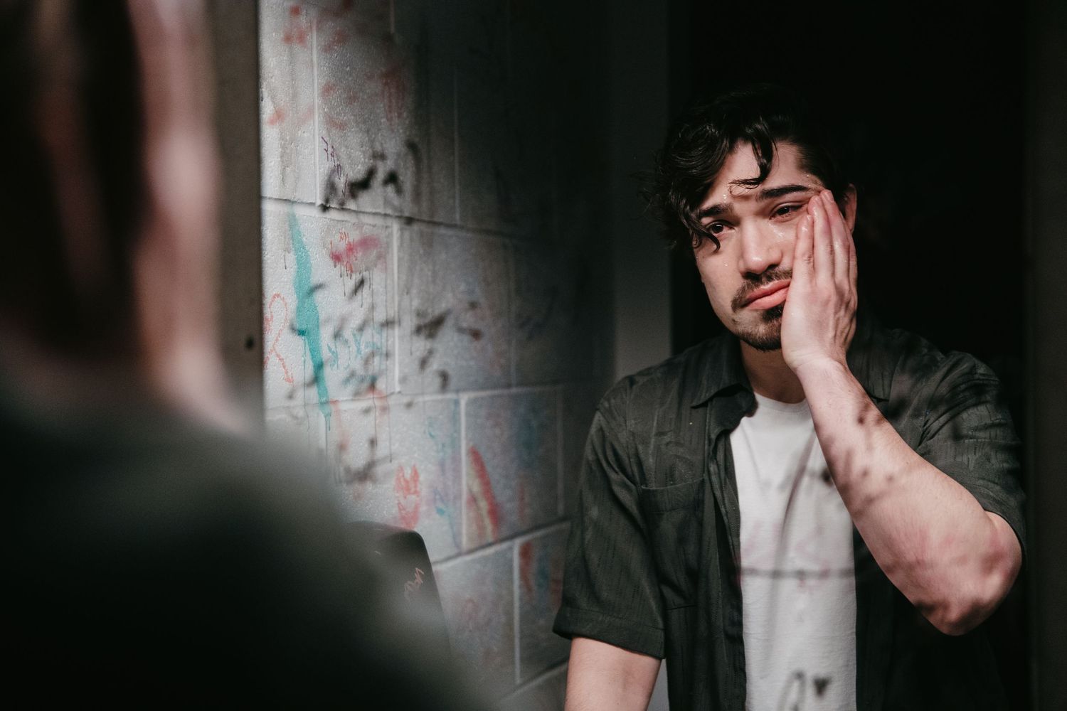 Young man with curly hair touching face while looking at reflection in graffiti bathroom mirror.