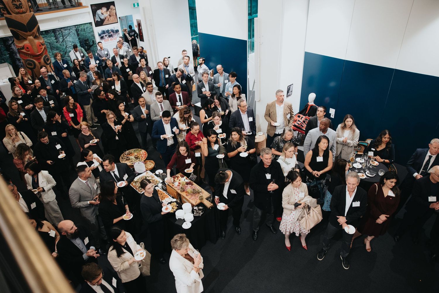 Large crowd gathered in indoor event space with white and teal walls.