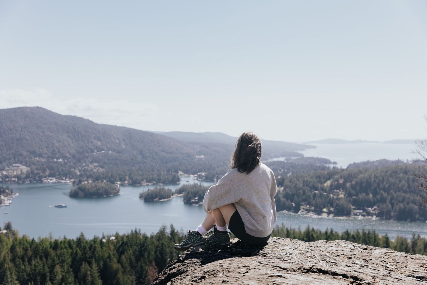 Person sitting on mountain peak overlooking lake and forest landscape.