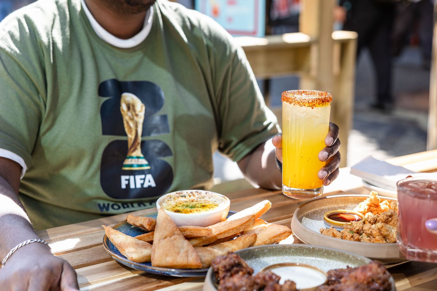 Person in FIFA World Cup shirt holding orange drink at table with hummus, pita, and appetizers outdoors.
