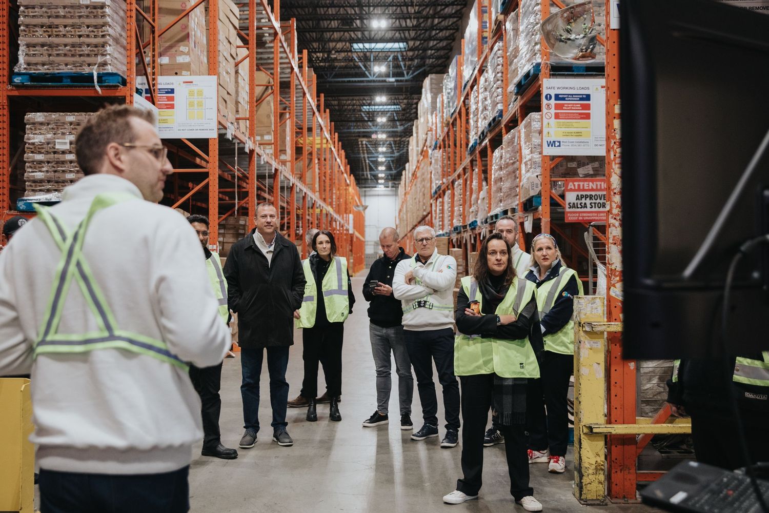Workers in high-visibility vests receive a warehouse tour among tall orange storage racks in a large distribution center.