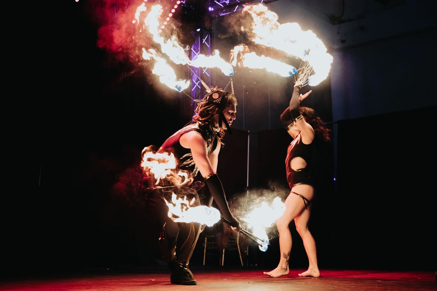 Two fire dancers perform a dramatic fire show on a dark stage with colorful smoke effects.