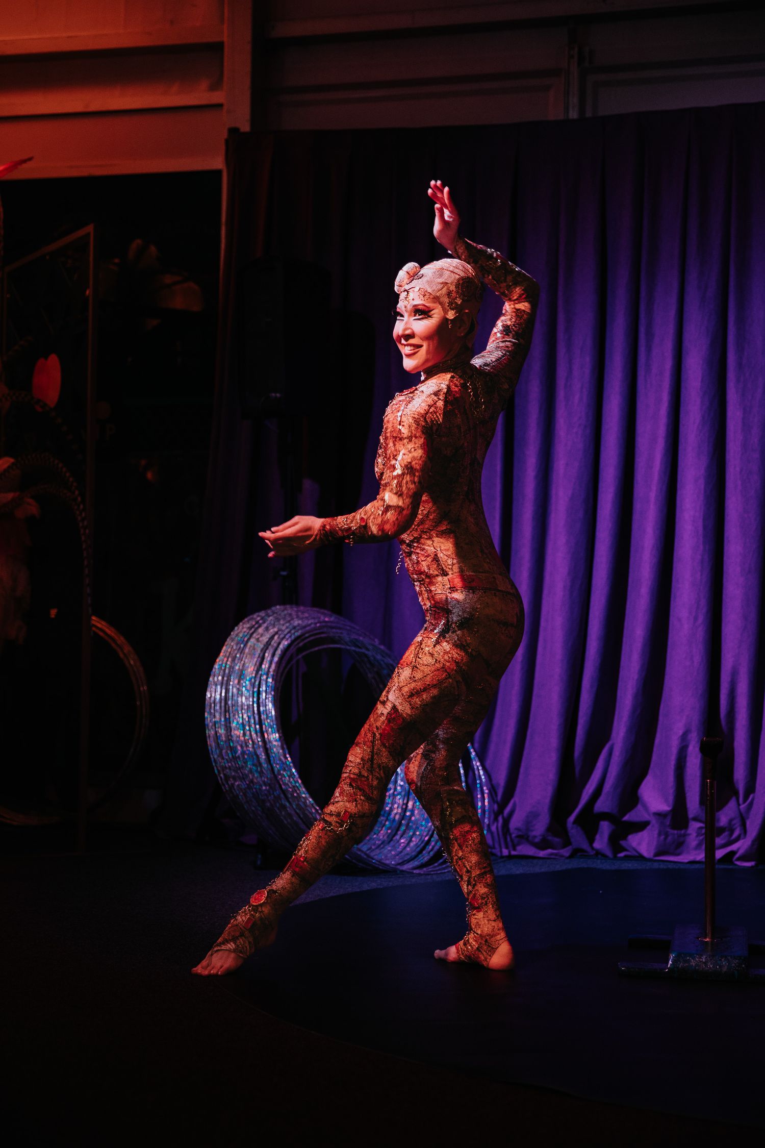 Circus artist in costume performs dynamic movements with metallic hoop on darkened stage.