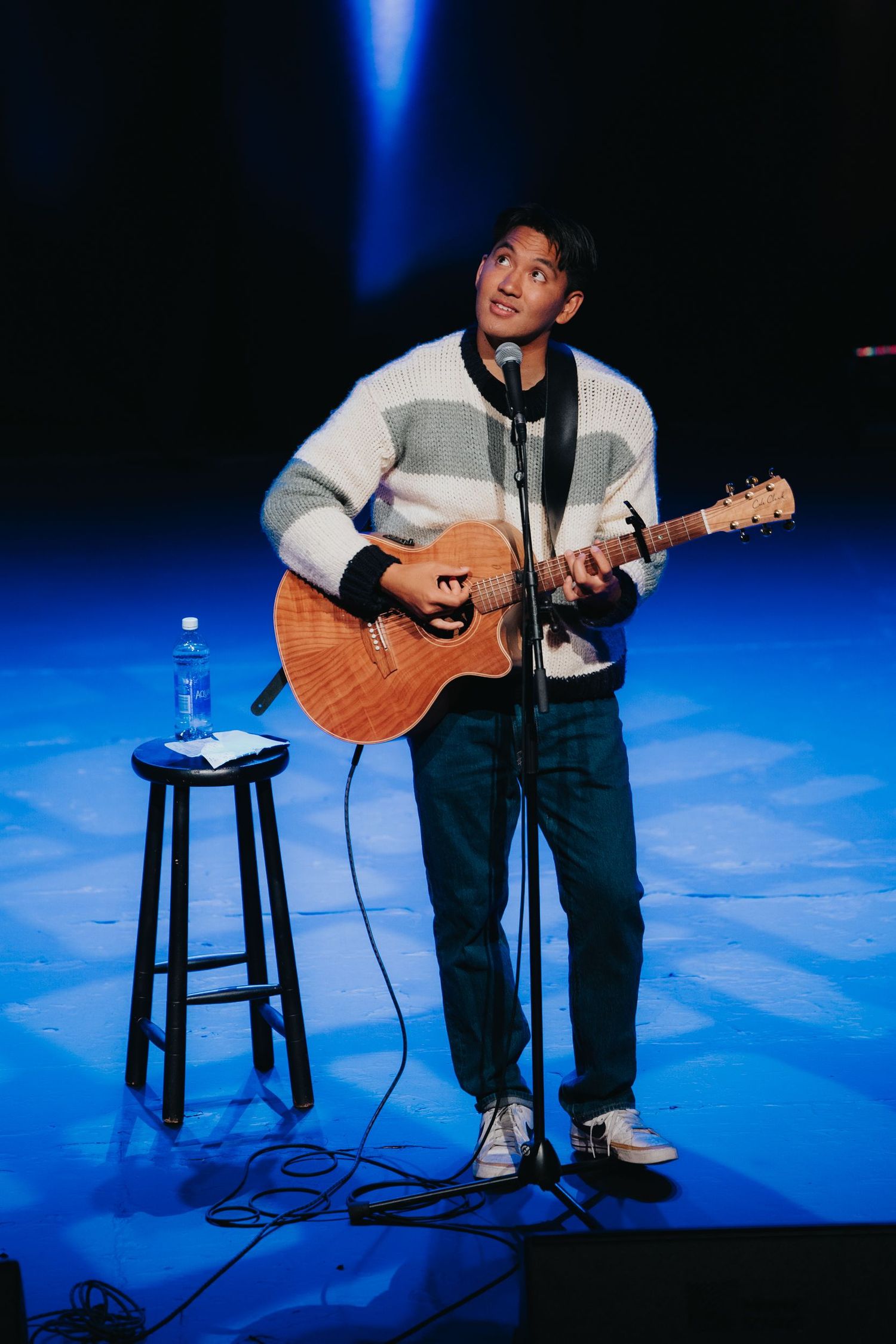 Young Asian man playing acoustic guitar on stage, wearing striped sweater and jeans, performing under blue spotlight.
