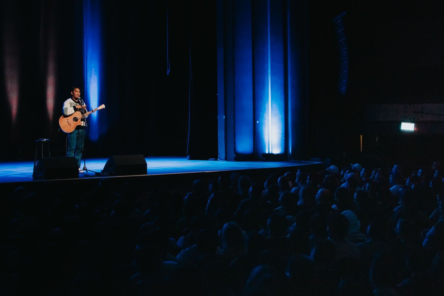 Solo guitarist performing on stage before large audience in blue-lit concert hall.