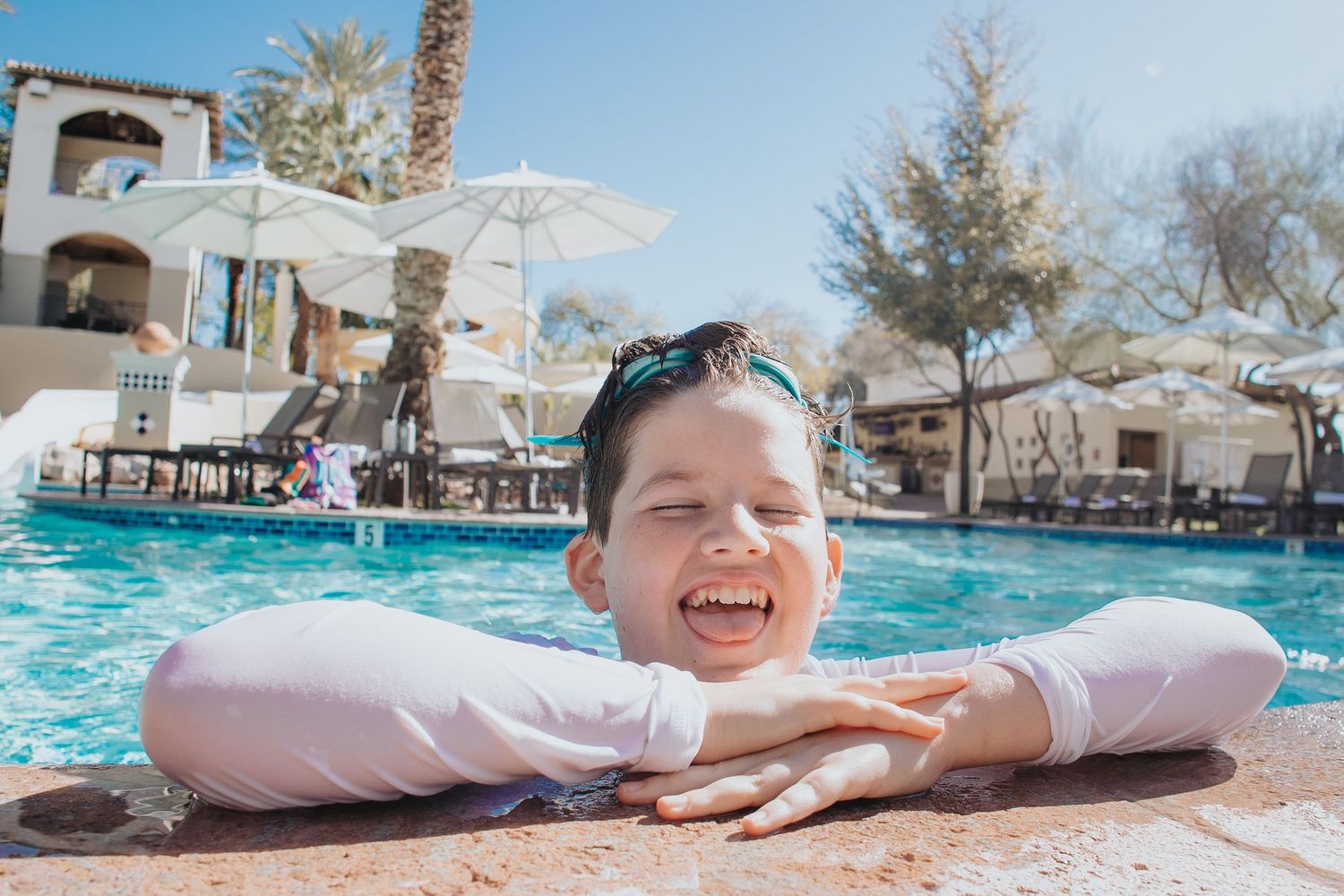People enjoying a sunny day at a resort pool with palm trees and lounge chairs.