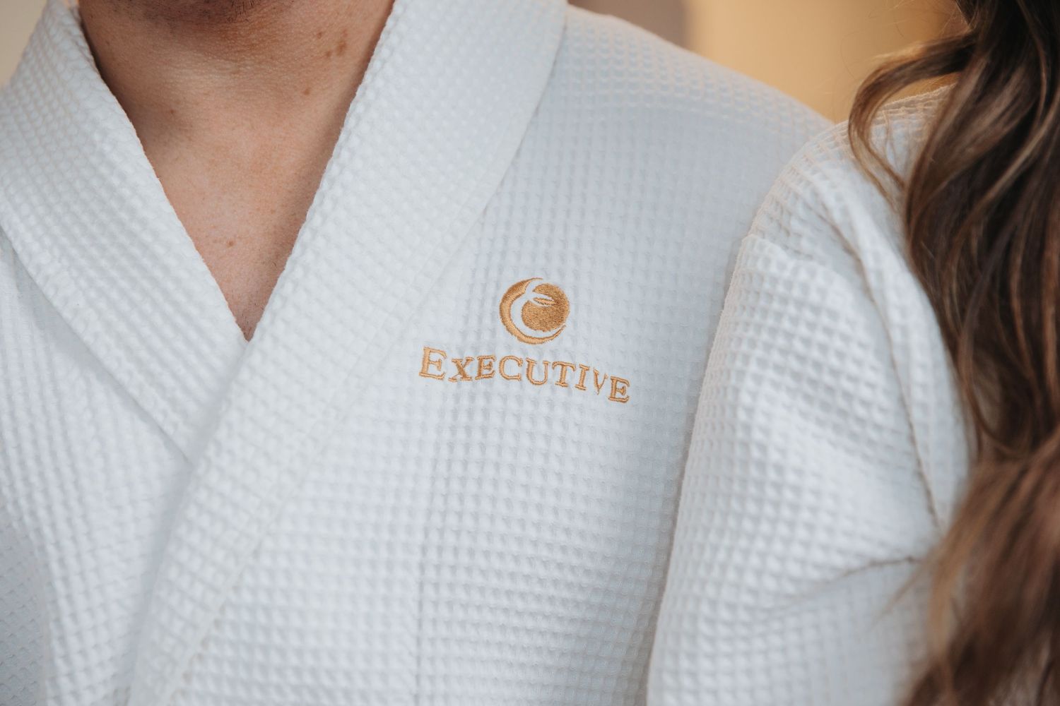 Close-up of a white waffle-knit Executive hotel spa robe with gold embroidered logo.
