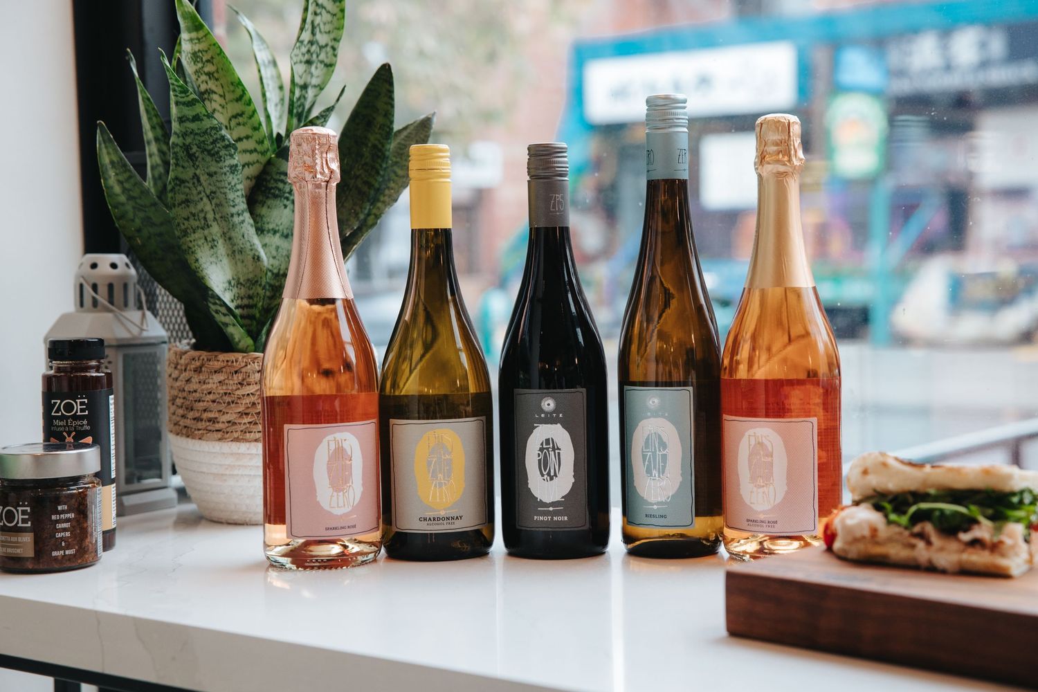 Collection of wine and spirit bottles arranged on a shelf with decorative plants in the background.