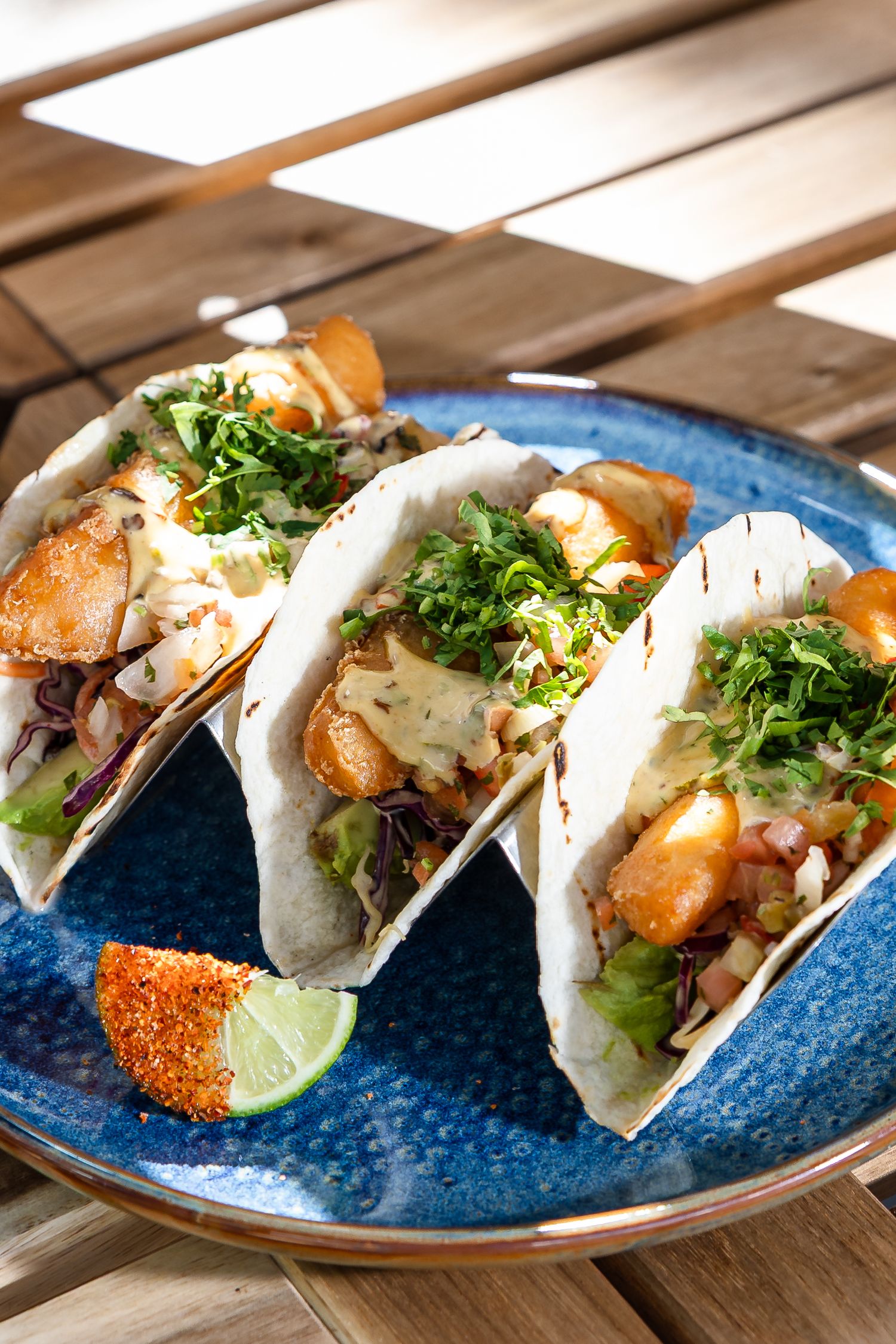 Three fish tacos with crispy battered fish, fresh herbs, and creamy sauce on a blue ceramic plate with lime wedge.