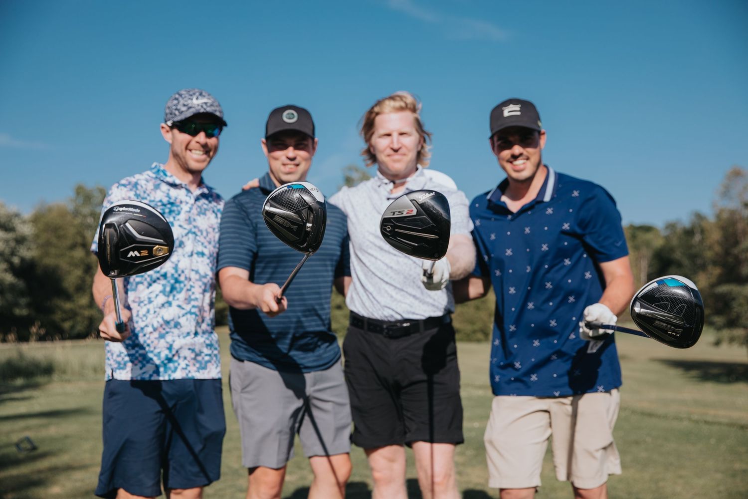 Four smiling men pose with golf clubs on a sunny golf course during a casual outdoor round of golf.