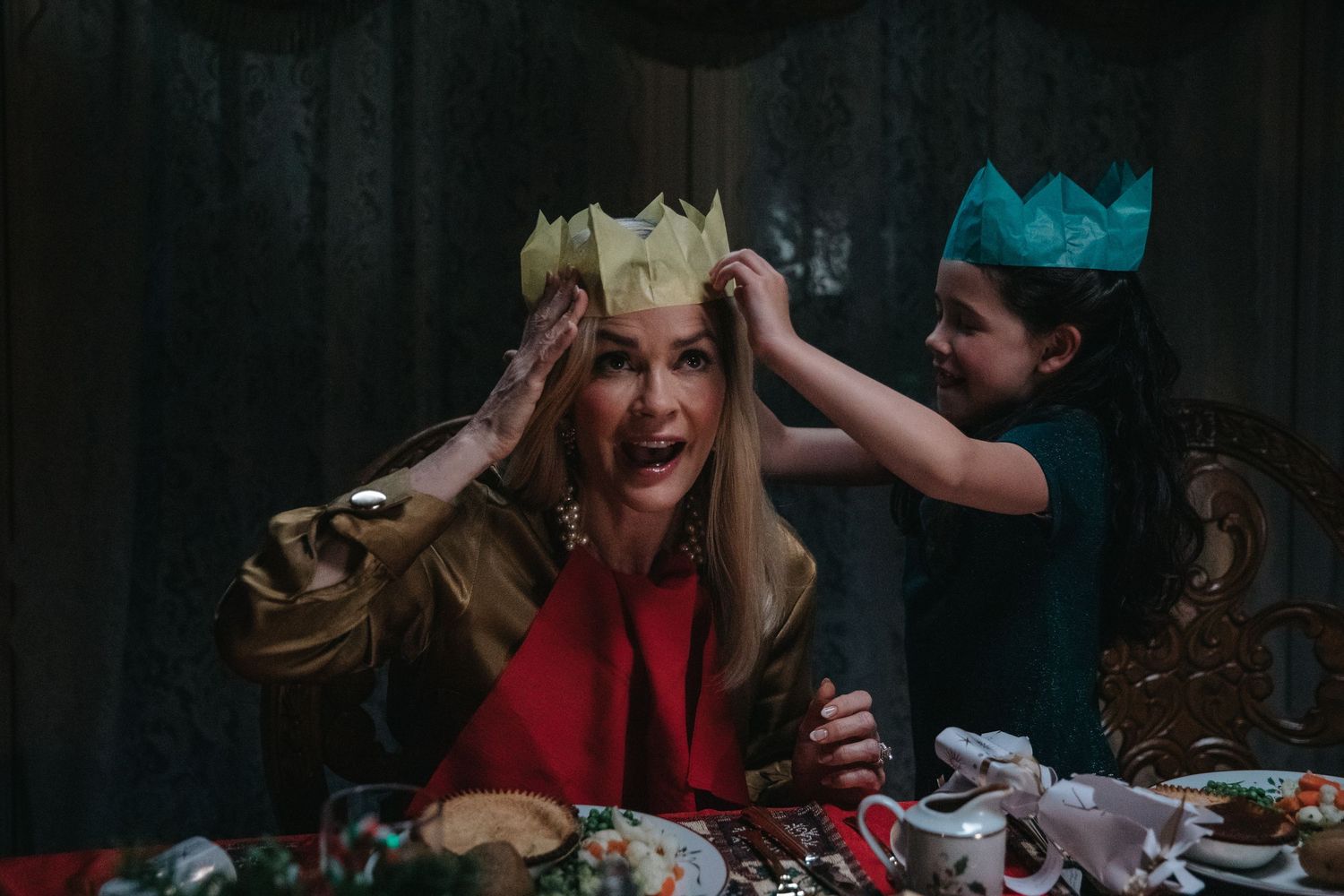 Woman in red dress laughs as girl places paper crown on her head at festive holiday table.