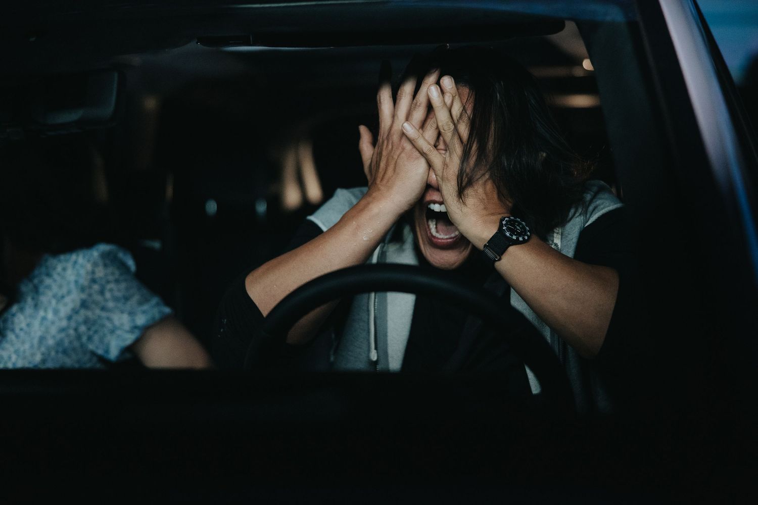 A distressed woman covers her face with both hands while sitting behind the steering wheel of a car at night.