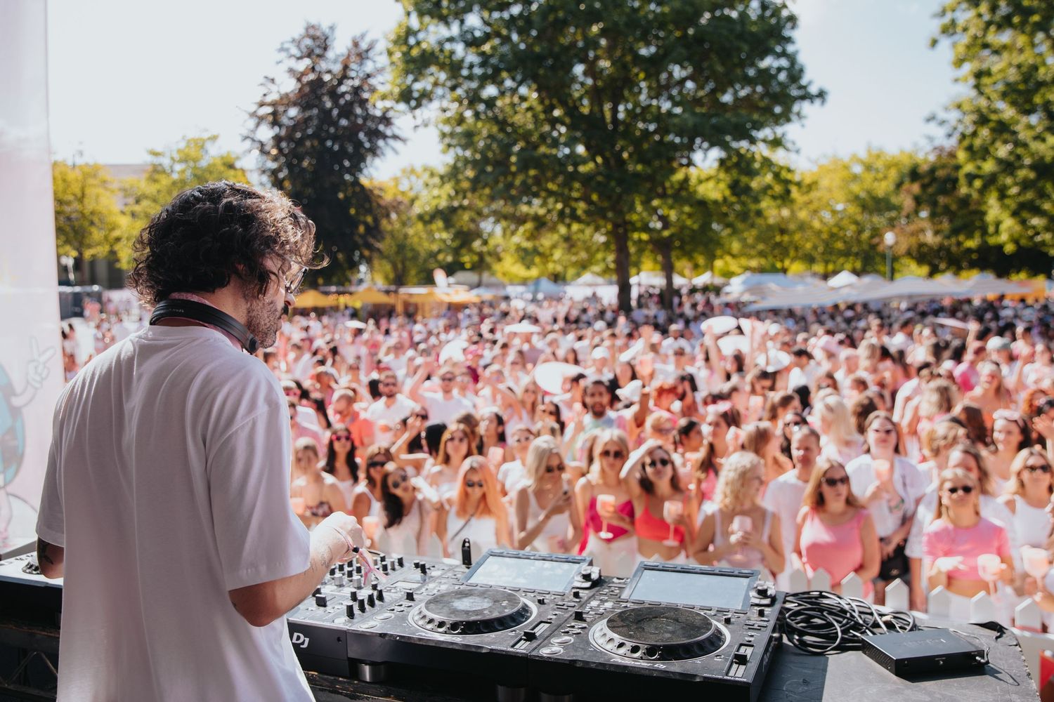 DJ performing at an outdoor summer festival for a large crowd of attendees dressed in white and pink attire.