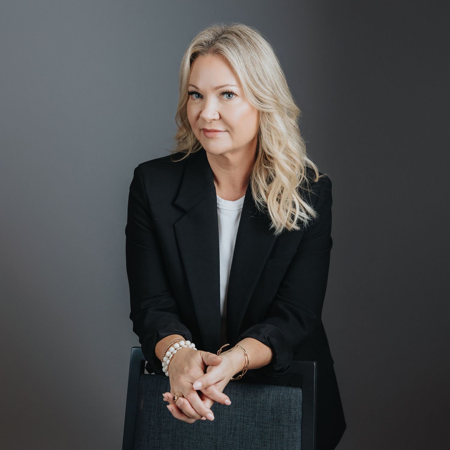 Professional woman in black blazer with blonde hair poses confidently with crossed hands in a corporate portrait.