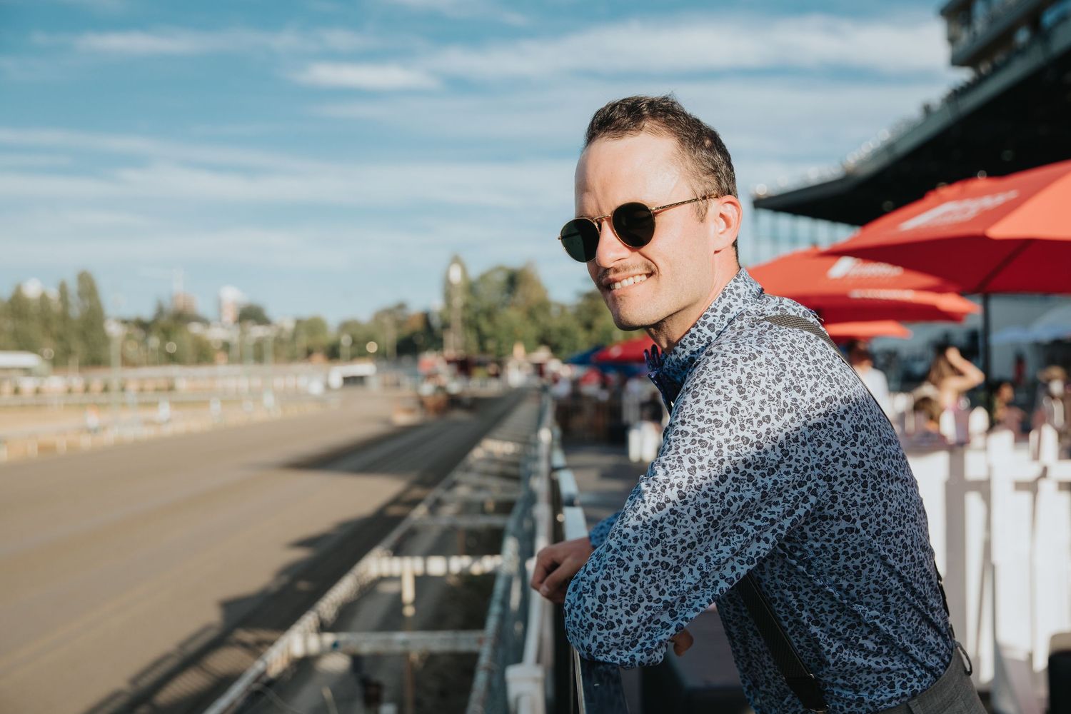 Smiling man in sunglasses and patterned shirt leans on railing at a sunny outdoor horse racing track.