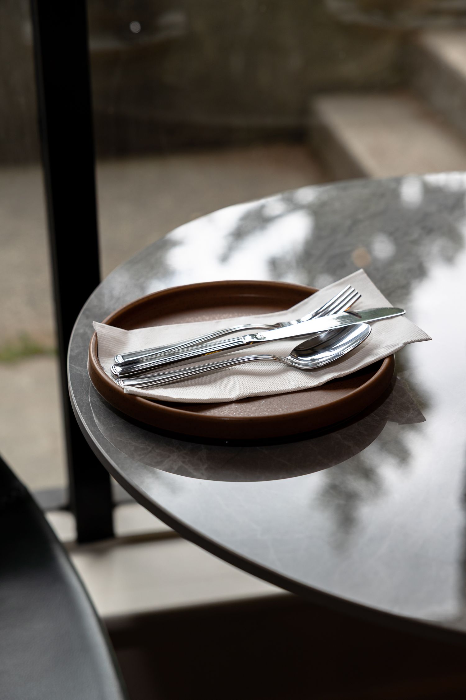 A rustic brown ceramic plate with silver fork, knife, and spoon on a napkin, placed on a round metal table.