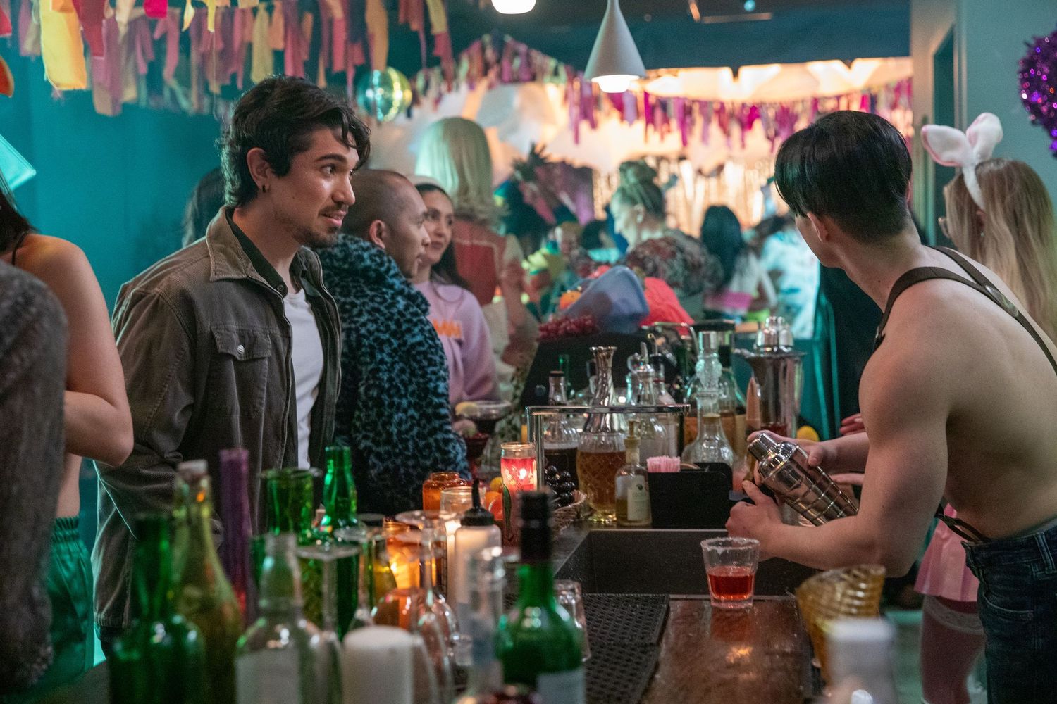 People socializing at a colorful bar party with bottles, candles, and festive decorations in the background.