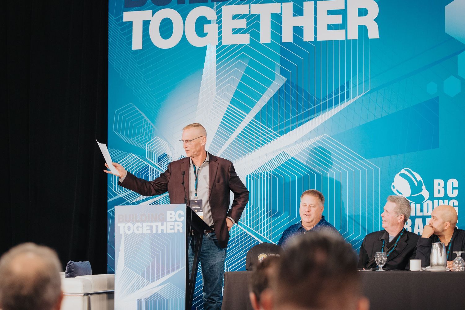 Speaker presenting at BC Building Trades Together conference, standing at podium before audience in modern event venue.