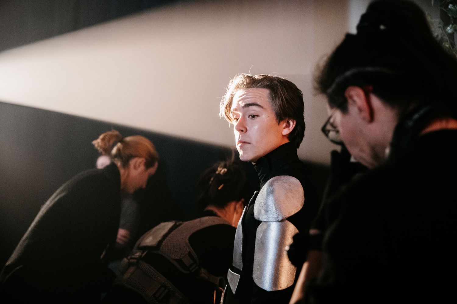 A young man in futuristic armor costume looks over his shoulder backstage at a dark theatrical production.