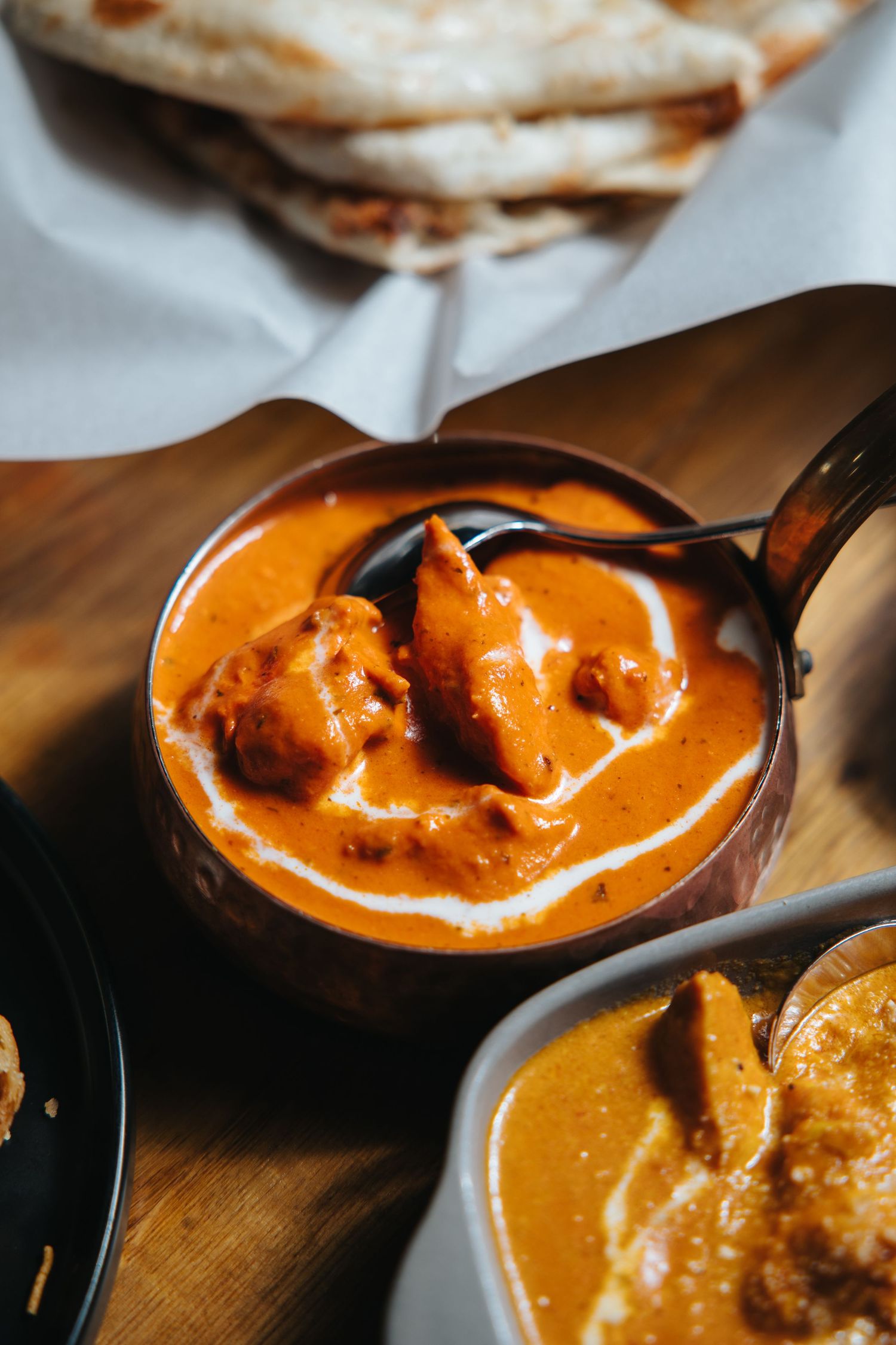 A copper bowl of creamy butter chicken curry with naan bread on a wooden board at an Indian restaurant.