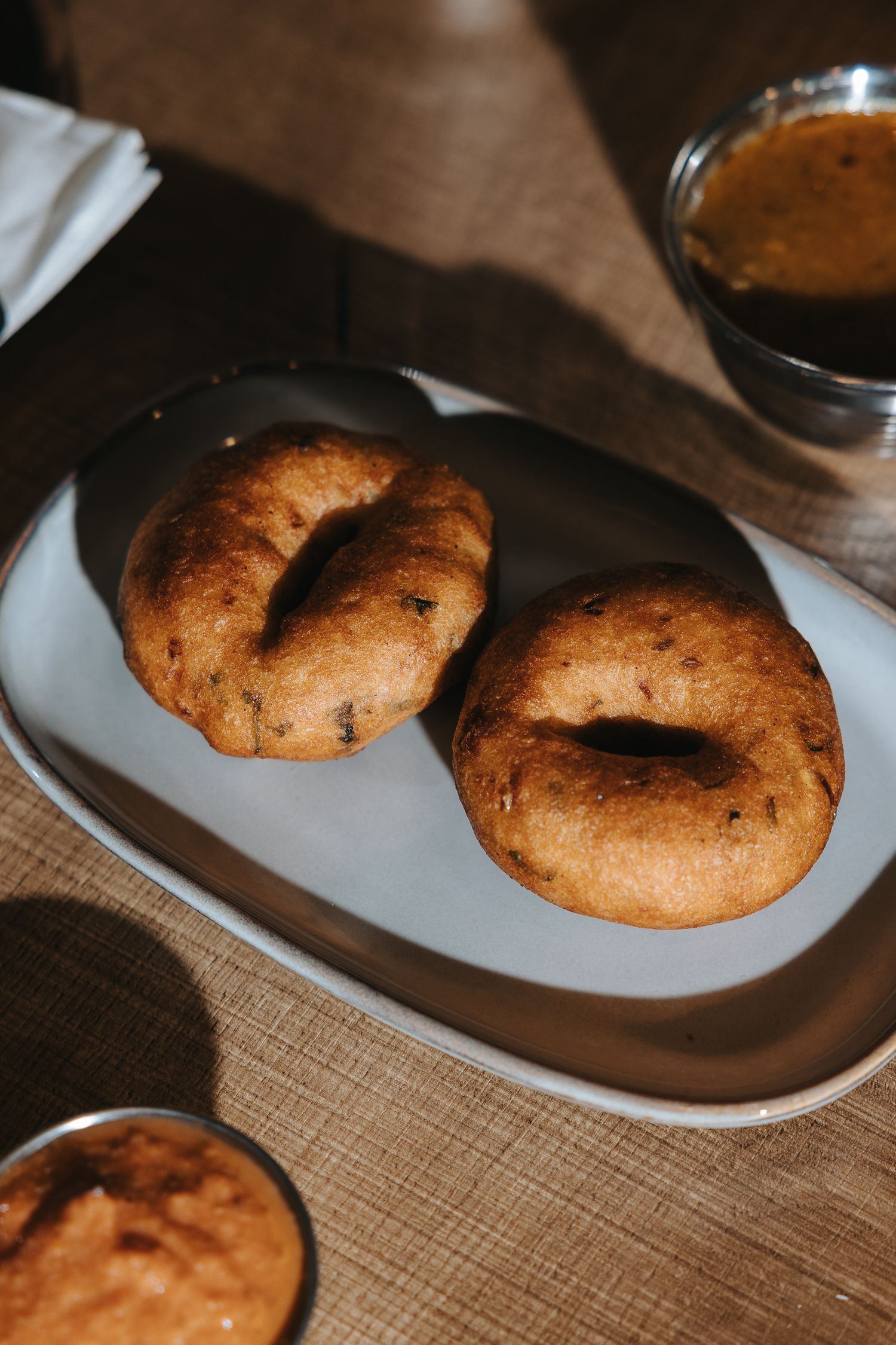 Two golden-brown crispy medu vada donuts served on a white plate with sambar dipping sauce on a wooden table.