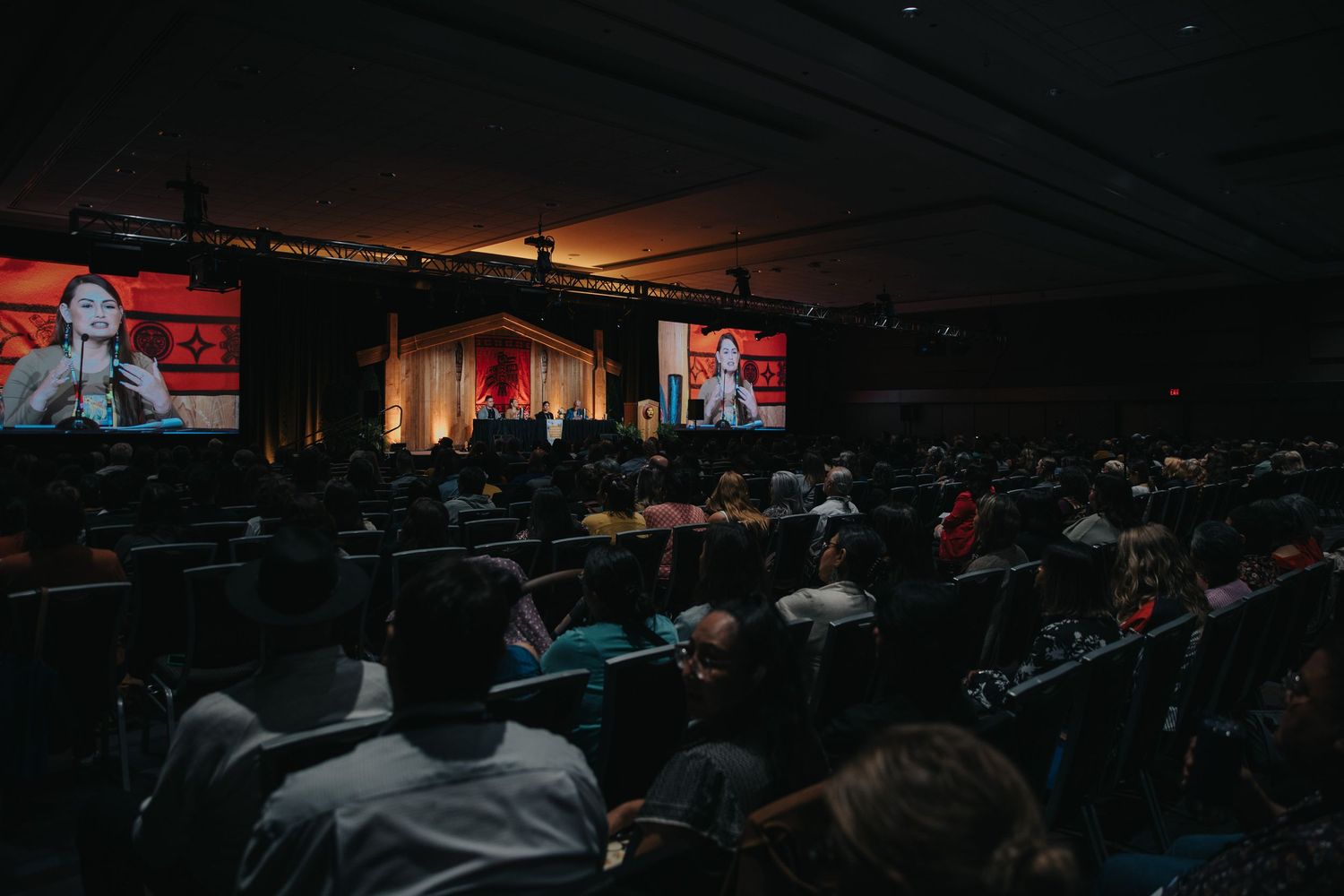 Dark auditorium filled with attendees watching presentation on large screens.