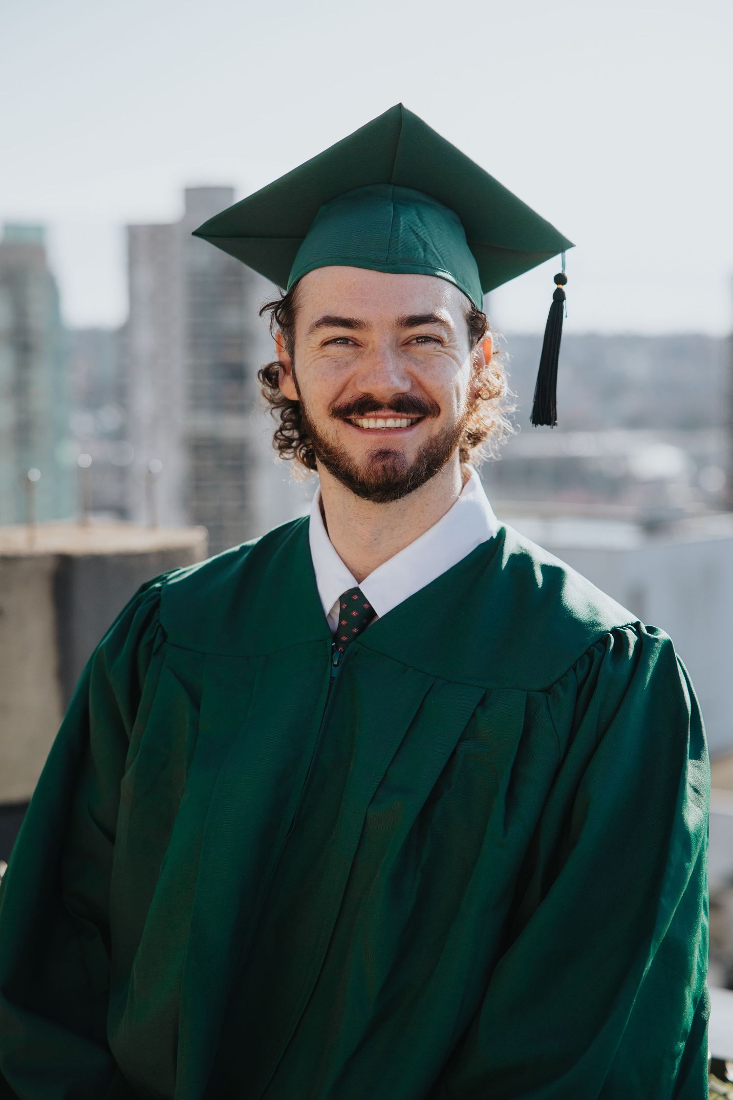 Graduate in dark green cap and gown smiling against city skyline backdrop.