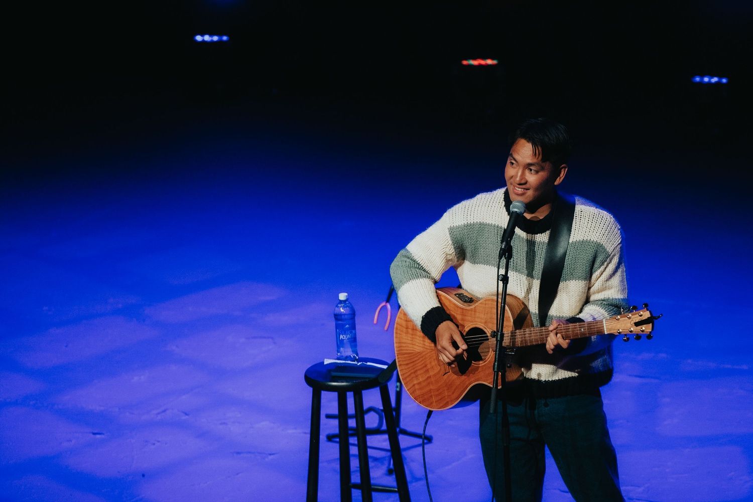 Musician playing acoustic guitar on stage under blue lighting with a stool and water bottle nearby.