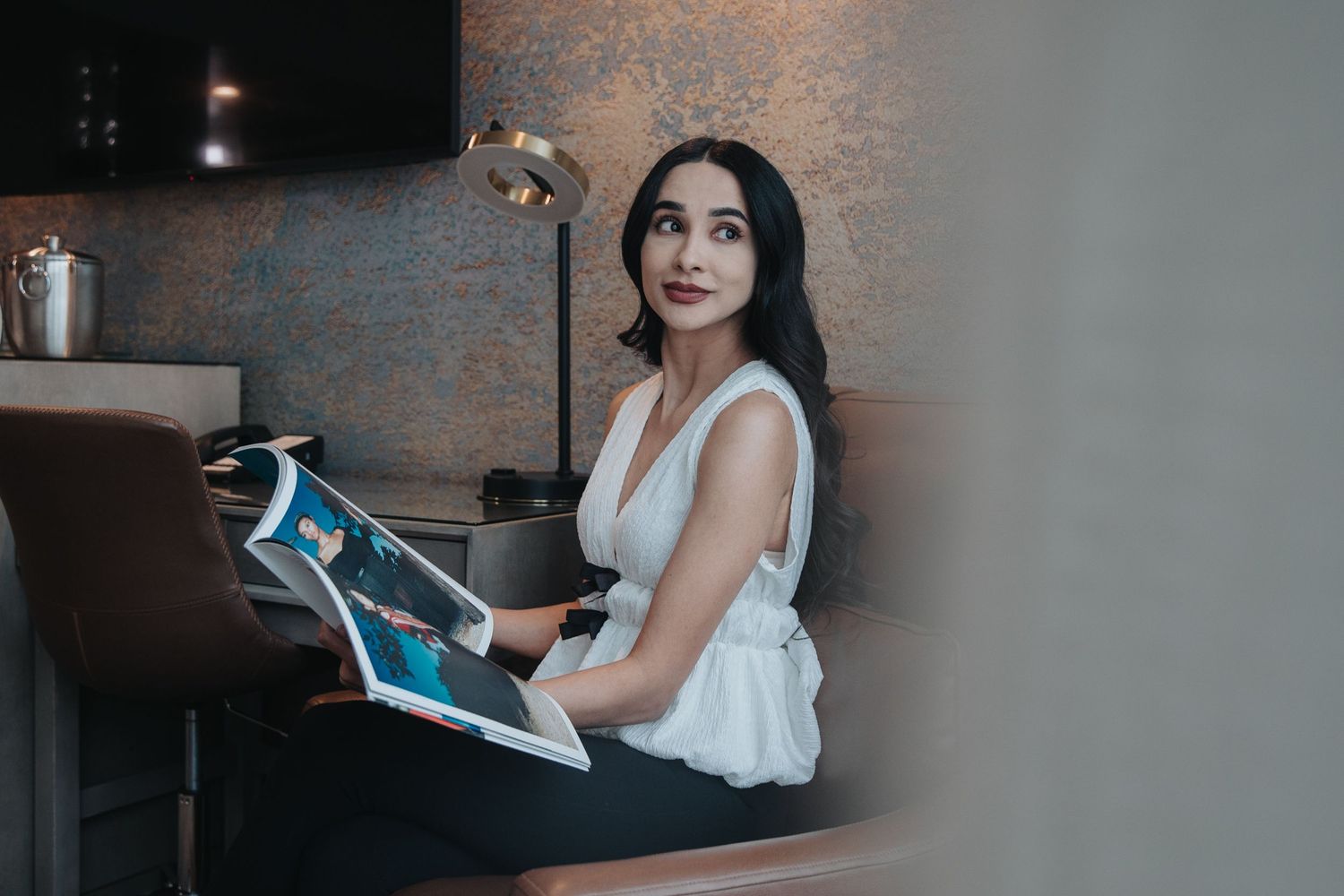 Young woman in white sleeveless top reading a magazine while seated at a modern desk with ambient lighting.