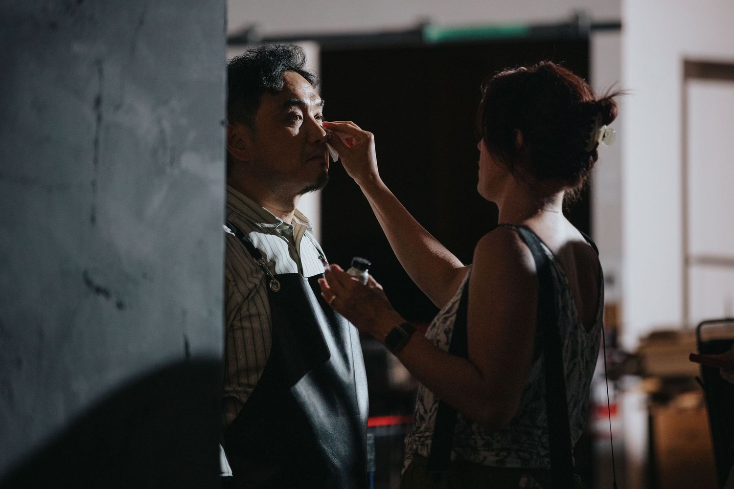 Makeup artist applying cosmetics to a man in a checkered shirt in a dimly lit studio setting.