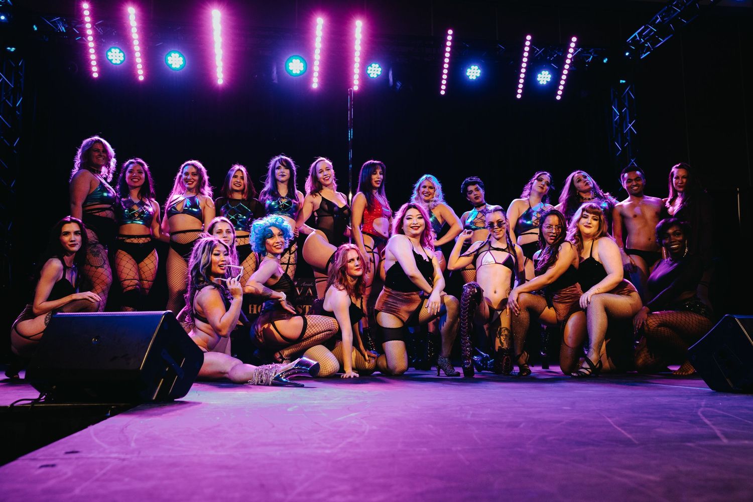 Diverse group of performers in lingerie posing together on a purple-lit stage after a burlesque show.