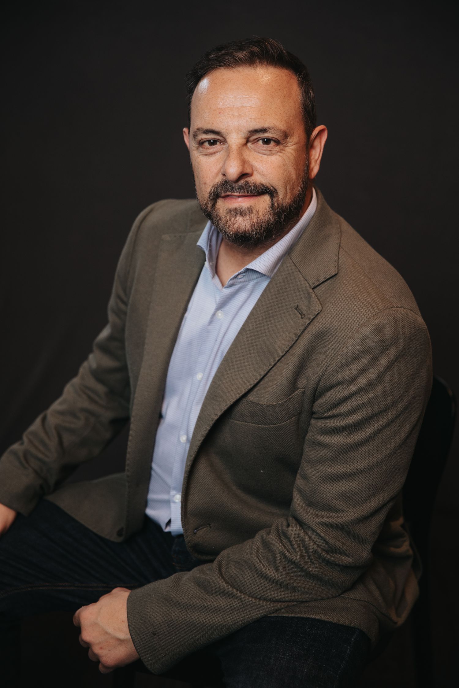 Business portrait of person in brown suit jacket and light blue shirt against dark background.