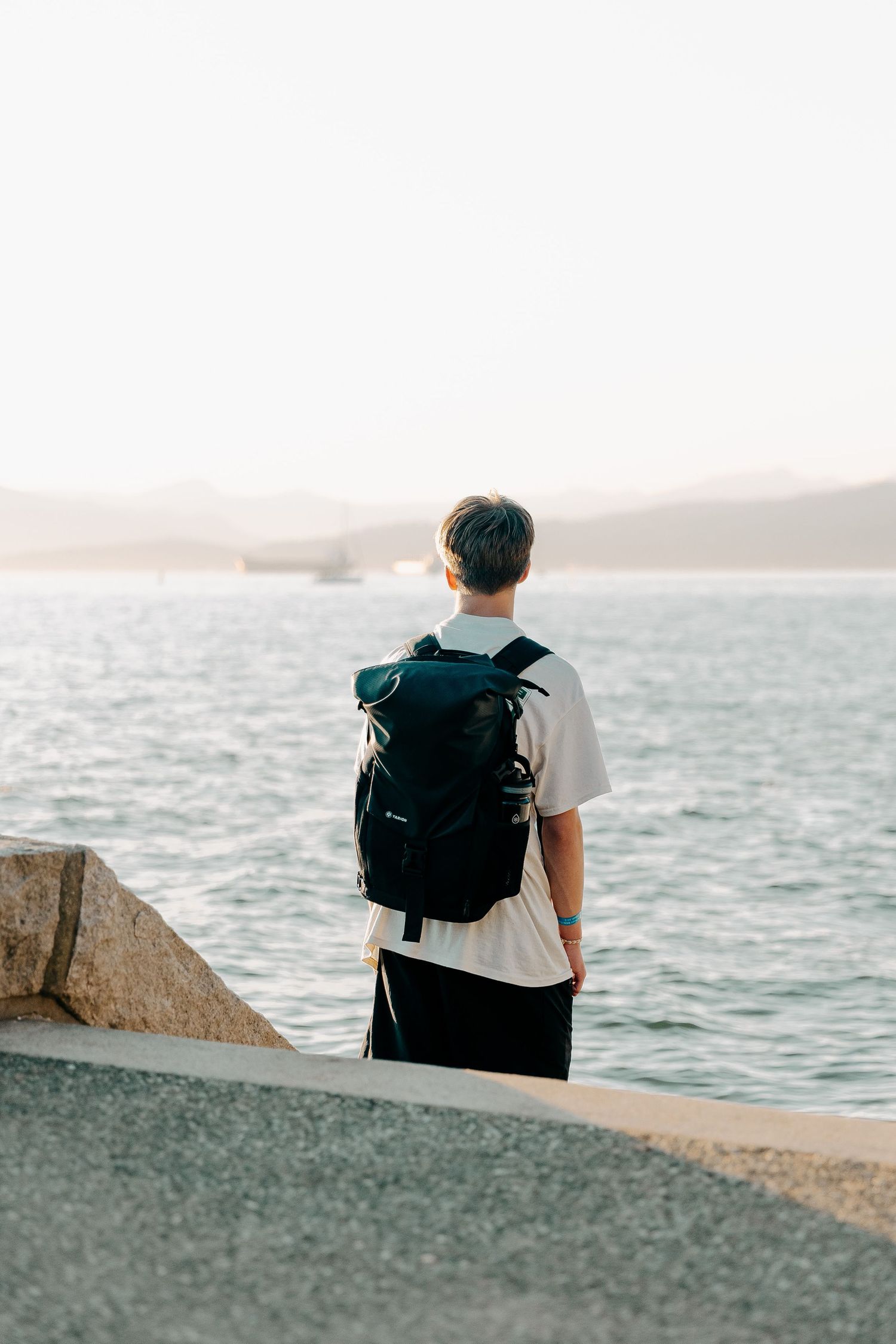 Person sitting on concrete barrier overlooking ocean view with backpack.