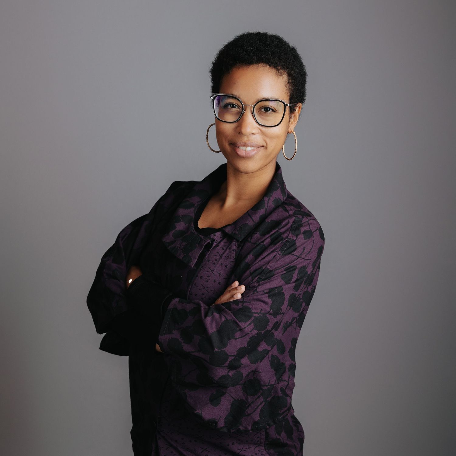 Person in glasses and purple patterned sweater poses against grey background in professional portrait series.