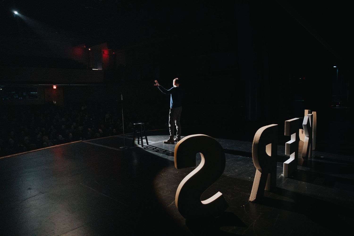 Performer standing on stage amid large illuminated letters before a packed concert crowd.