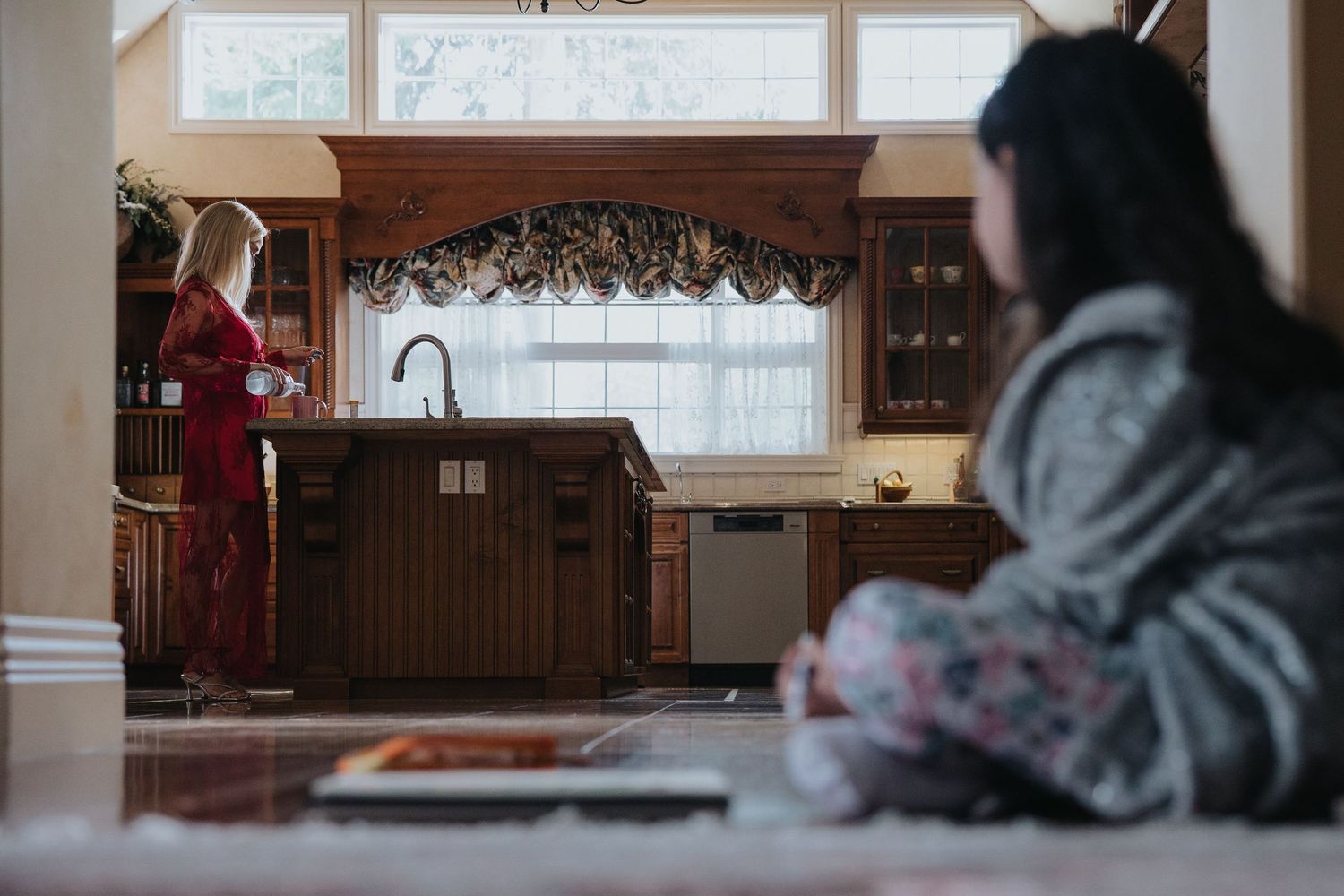Woman in red robe stands at kitchen sink while child sits in foreground of elegant wood-paneled kitchen.